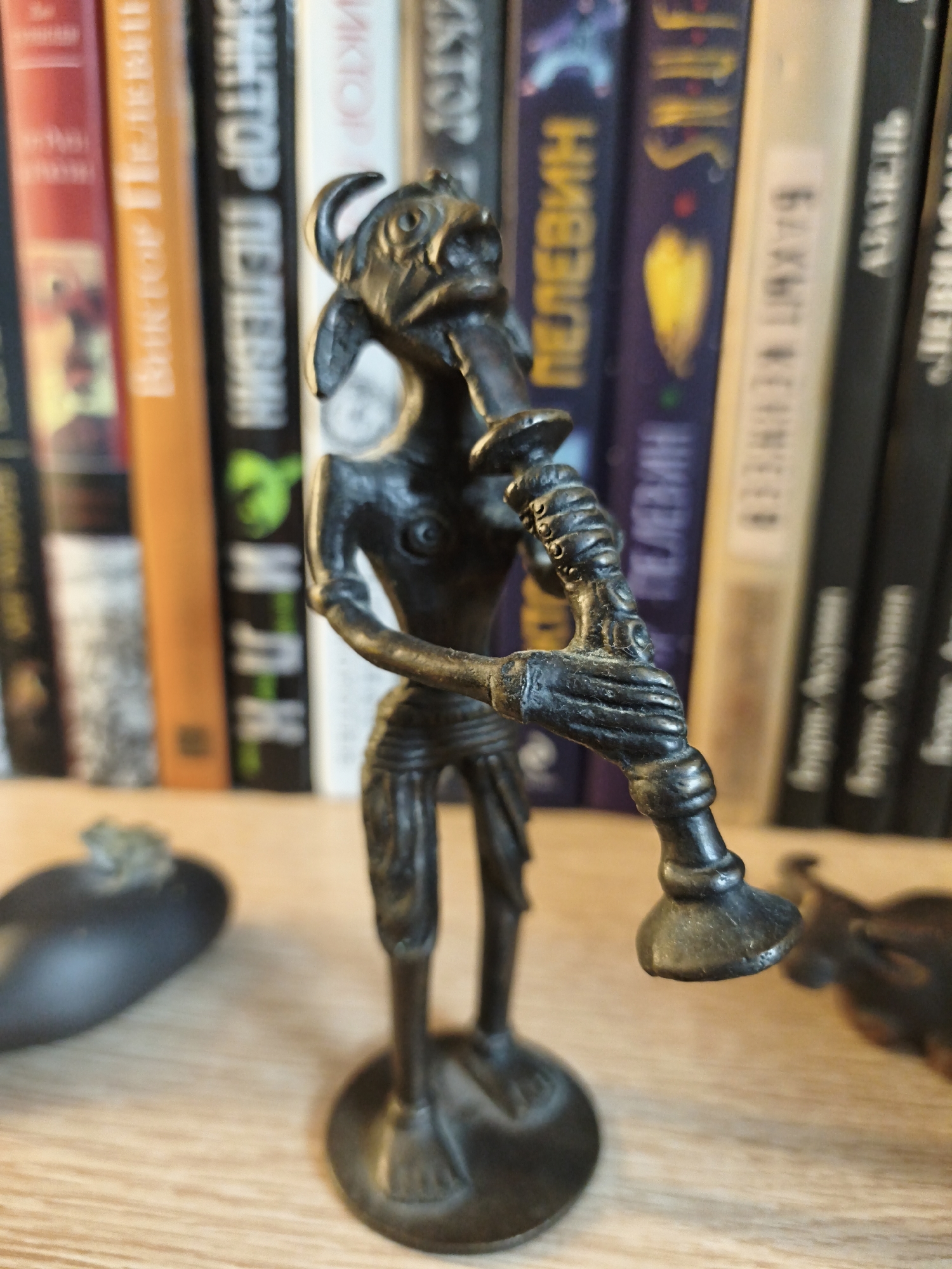 This image captures a close-up of a dark, metallic figurine of a creature playing a wind instrument, possibly a flute or pipe. The figurine is depicted in a standing, somewhat hunched posture, with exaggerated features and a skeletal appearance. It stands on a circular base. In the background, a shelf is filled with books, suggesting an indoor setting, likely a home or library. The books are varied in color, with some spines displaying text in a non-Latin script, possibly Cyrillic, which could indicate the location. There are also other objects on the shelf, including what appears to be an eggplant-shaped object with a small green element on top to the left of the figurine, and a blurred, dark object to the right. The lighting is diffused, casting soft shadows, and the depth of field blurs the background, drawing attention to the intricate details of the figurine. No specific time of day or weather conditions are discernible. The overall mood is somewhat mysterious and artistic. The text on some of the book spines is not clearly legible, but some characters resemble Cyrillic letters. The location context provided is Astana, Kazakhstan, which aligns with the potential presence of Cyrillic text on the books.