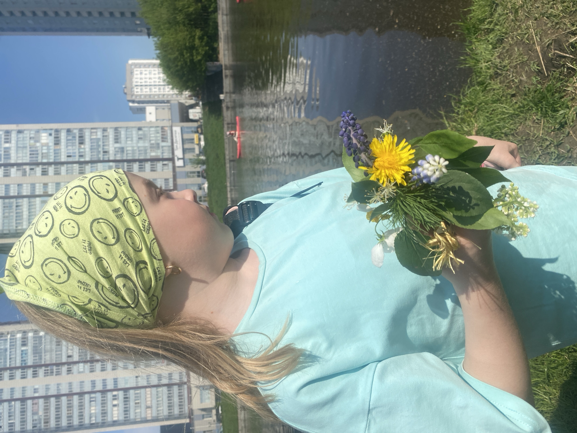 A young person with blonde hair and a green smiley face bandana stands outside with a bouquet of wildflowers. The person is wearing a light blue t-shirt. In the background, there is a body of water and high-rise buildings, suggesting an urban environment. A red kayak is visible on the water. The location is likely the city of Yekaterinburg, Russia.