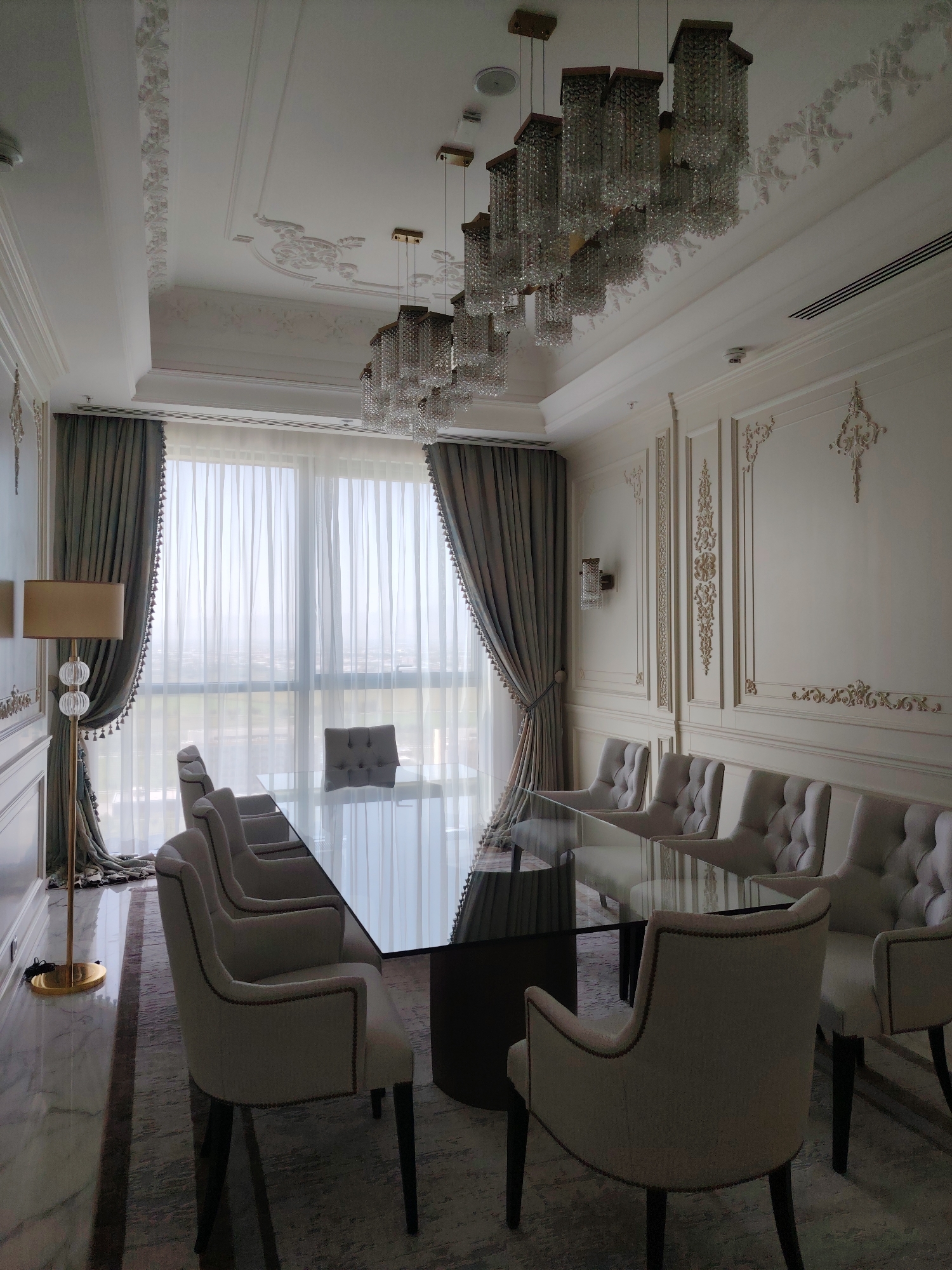 In a formal dining room setting in Konigil, Uzbekistan, a large, rectangular glass-topped dining table is the central focus, reflecting the ornate crystal chandeliers suspended from the intricately decorated ceiling. Around the table, several upholstered dining chairs with high backs and button-tufted detailing are arranged, suggesting a capacity for multiple diners. The room is decorated in a luxurious style, with detailed molding on the walls and ceiling, and heavy, draped curtains framing large windows that offer a view of a hazy, possibly urban, landscape. A floor lamp with a metallic base and a fabric shade stands near the window, adding to the room's sophisticated ambiance. The flooring consists of polished marble tiles with a patterned rug beneath the table and chairs. The overall impression is one of opulence and formal elegance.