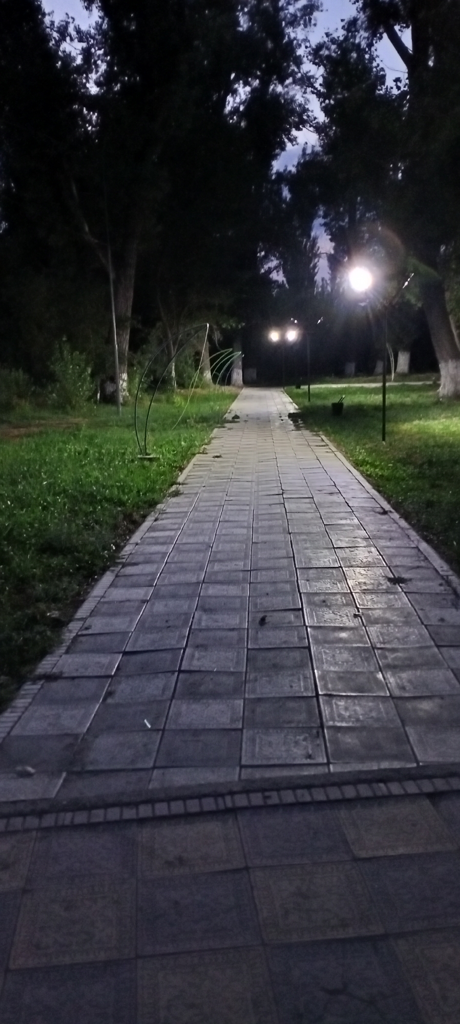 This is a medium-shot, vertical, eye-level, photographic image of a paved walkway in a park-like setting, likely during twilight. The walkway, composed of square gray pavers with intricate patterns, recedes into the background, creating a sense of depth. To the left of the walkway is a grassy area with some scattered debris. Several trees with dark foliage line the sides of the path, and some are illuminated by streetlights. In the distance, there are two glowing streetlights, and a third, brighter streetlight is visible to the right, casting a glow on the wet pavement. The sky above is a muted twilight blue, with darker tree branches silhouetted against it. The overall impression is one of quiet solitude in a park setting at dusk. The location is identified as Sokuluk, Kyrgyzstan, suggesting this is a public park or boulevard within the town. There are no people visible in the image.
