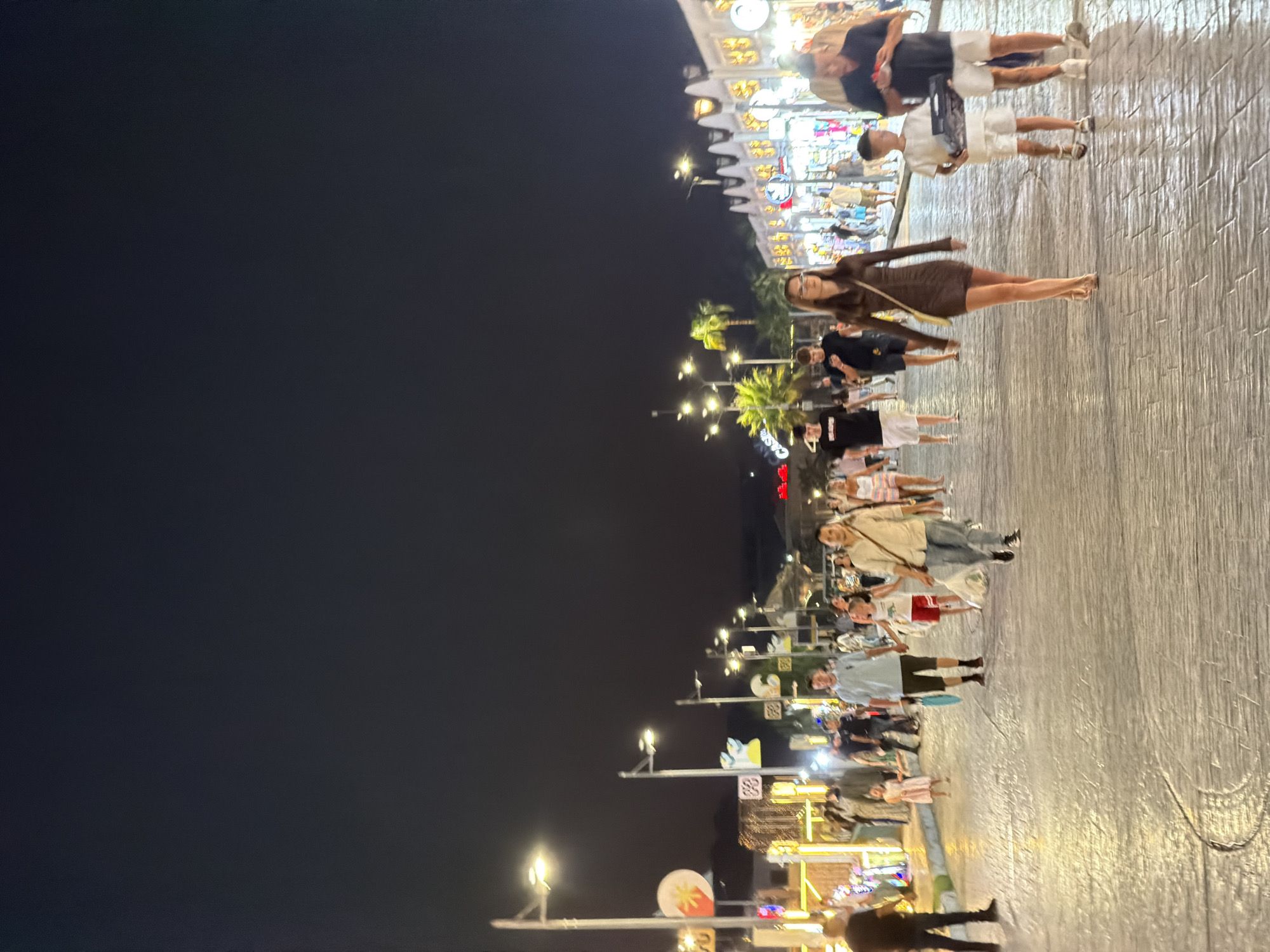 The image shows a street scene at night in Sharm El Sheikh, Egypt. The street is paved and lined with shops and businesses that are illuminated by lights. People are walking along the street, some individually and some in groups. The sky is dark, indicating that it is nighttime. The overall impression is one of a busy and vibrant urban environment.