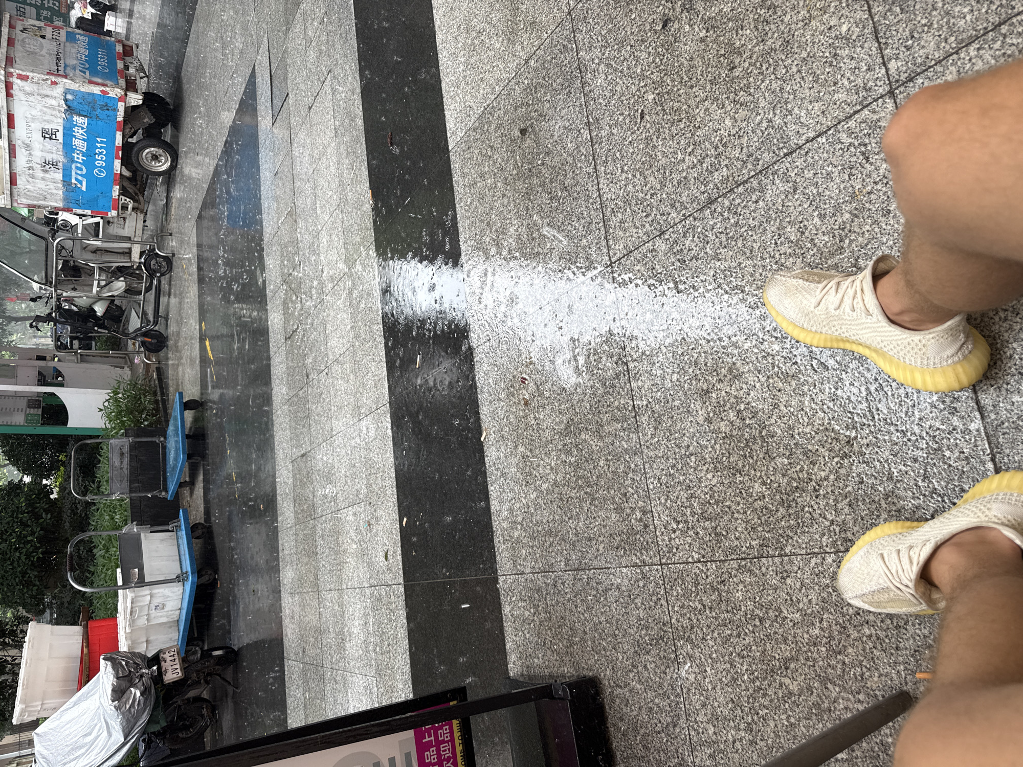 The image shows a close-up view of a person's legs and feet wearing light-colored sneakers with yellow soles, standing on a wet, speckled granite tile floor. A thin, white line of spilled powder or liquid runs across the floor from the left side of the frame, disappearing to the right.
In the background, to the left, a delivery truck with "中通快递" (ZTO Express) branding is partially visible. The truck is white with blue and red accents and features the phone number "95311". Next to the truck are several blue and black wheeled carts, some carrying white boxes. A motorcycle with a license plate "JV1442" is also parked nearby, partially covered by a silver tarp.
The ground appears wet, suggesting it might be raining or has recently rained, indicated by ripples in the water and reflections on the dark tiles. The overall scene suggests an urban setting, likely a sidewalk or loading area outside a business, given the delivery truck and carts. The lighting and reflections hint at a daytime scene. Visible text on the truck and license plate provides specific identifying information. The image captures a moment in time related to logistics and delivery operations.