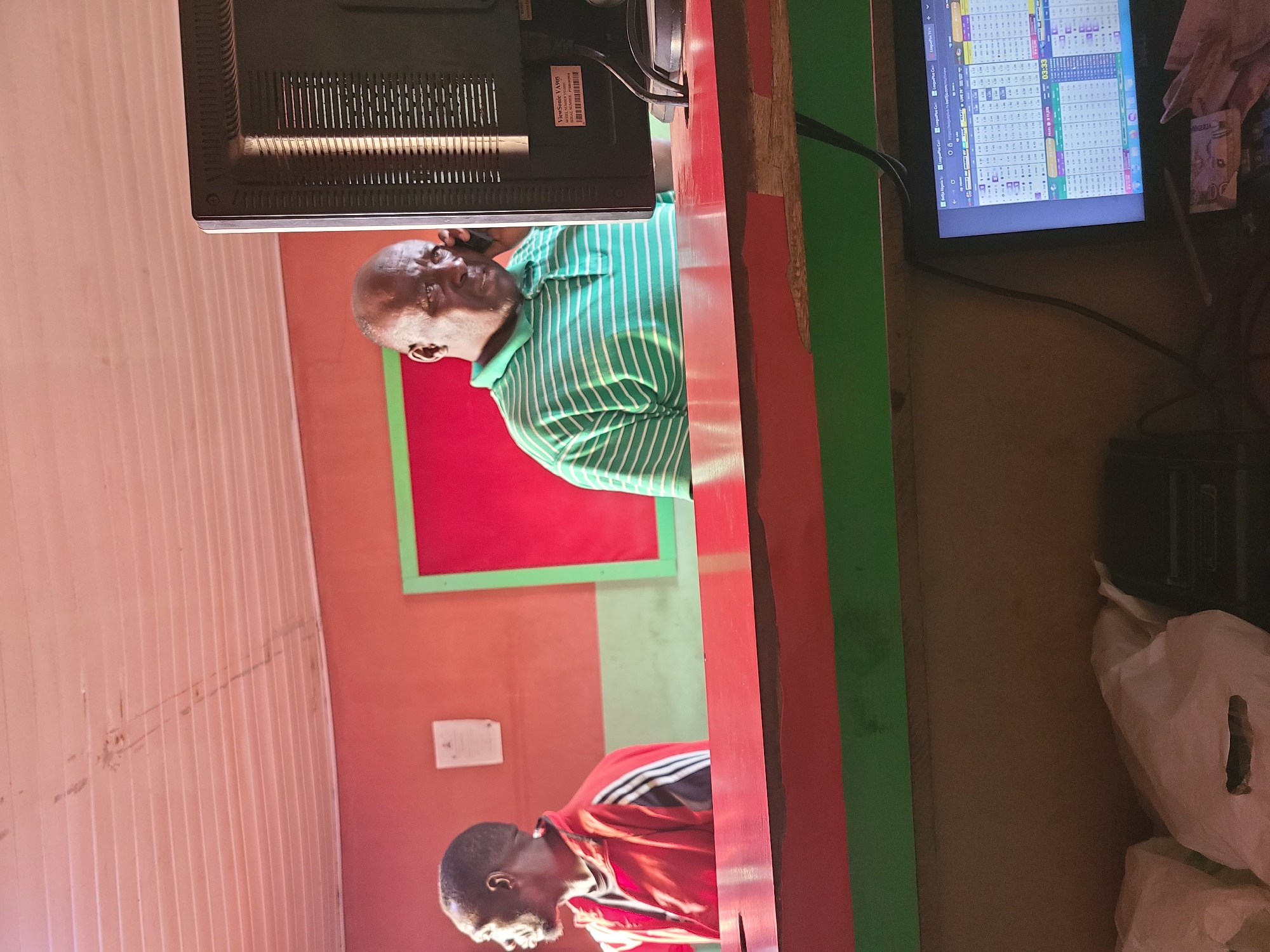 This indoor image features two men engaged in an activity, likely within a small business or betting shop. The primary subject is a bald man wearing a green and white striped polo shirt, seen from the chest up. He is holding a mobile phone to his right ear, seemingly in conversation. He is positioned behind a bright red counter, which also features a green section.
Behind the man in green, a computer monitor is visible, its back displaying a label that reads "ViewSonic VA905." To the right of this, another computer monitor is active, showing a grid of numbers and text. Visible text on the screen includes "Betja Nigeria," "LonglegsPC.com," "LIVE STATS," and a digital timer set to "03:33." This content strongly suggests an online sports betting platform or live score display. A crumpled Nigerian Naira note is also visible near this monitor, resting on the counter.
Further into the scene, another man is visible, wearing a red and black long-sleeved top. He is positioned to the left (when correctly oriented) of the man in green, also behind the counter, and appears to be looking downwards.
The background consists of distinct colored sections: a large red wall with a bright green frame behind the man in green, and a textured, light peach-colored corrugated wall further to the left. A small, white rectangular object is affixed to the red wall. On the far right side of the image, near the second monitor, several white plastic bags are visible, suggesting stored items or waste.
The scene depicts a functional, brightly colored commercial space, with the men focused on their respective tasks. The visible text and currency note reinforce the location context of Jos, Nigeria, and indicate a business operation likely involving betting or digital services. The general lighting suggests it is daytime.