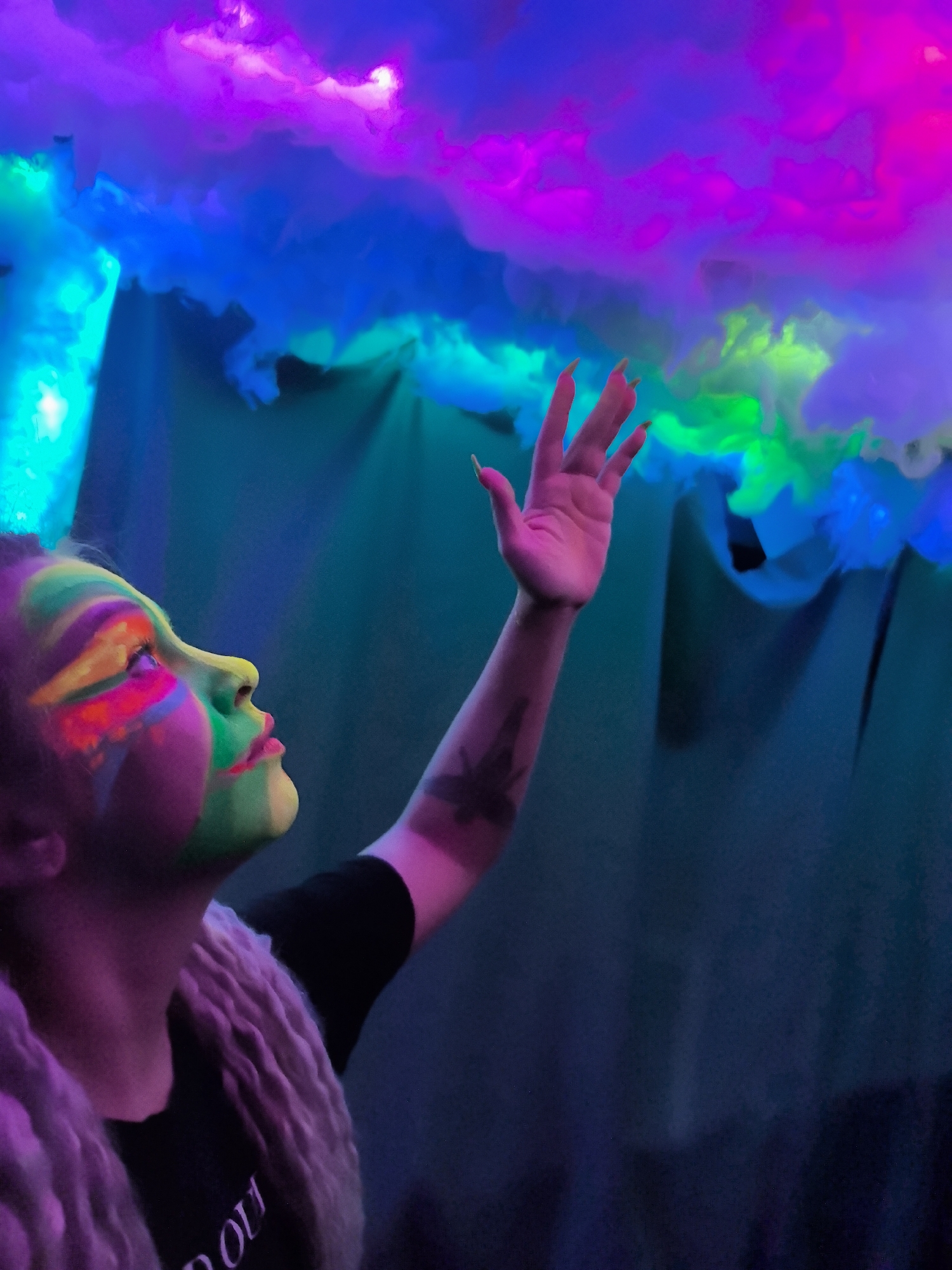 A person with vibrant face paint, illuminated in a dark room, reaches upward with an outstretched hand toward colorful, cloud-like structures suspended above. The face paint consists of a rainbow array of colors, including yellow, green, pink, and orange. The person is wearing a black t-shirt with indiscernible text and a light-colored fuzzy vest. The cloud structures are illuminated with varying colors like purple, blue, and green. The backdrop appears to be a dark fabric. There is no evidence that allows us to determine if the scene is located in the city of Noviy, Russia.