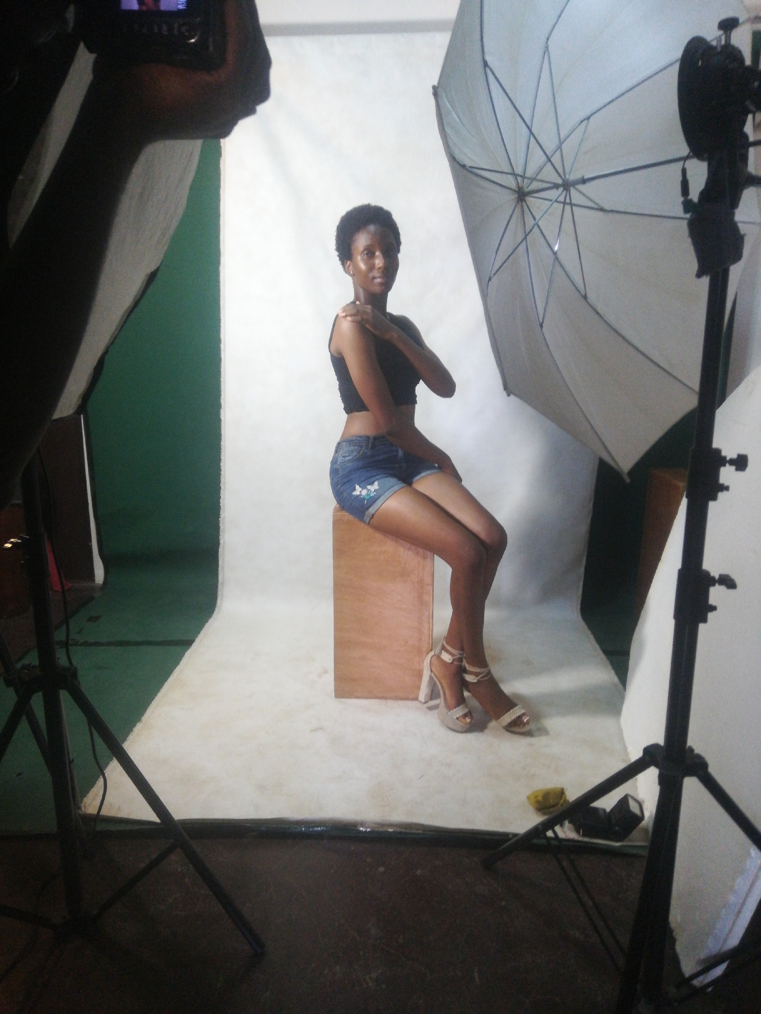 In an indoor studio setting, a young woman with dark, short hair is posing for a photograph. She is seated on a wooden cube, wearing a black tank top and denim shorts with white floral embroidery. Her legs are crossed, and she is wearing strappy white heeled sandals. A large, octagonal white umbrella is positioned behind her, angled downwards. Lighting equipment, including softboxes and a tripod with a flash, surrounds the subject, indicating a professional photoshoot. The backdrop is a seamless white paper, with a green wall visible behind it. The floor is dark and appears to be made of wood or linoleum. The image captures a moment in a controlled studio environment, likely for fashion or portrait photography.