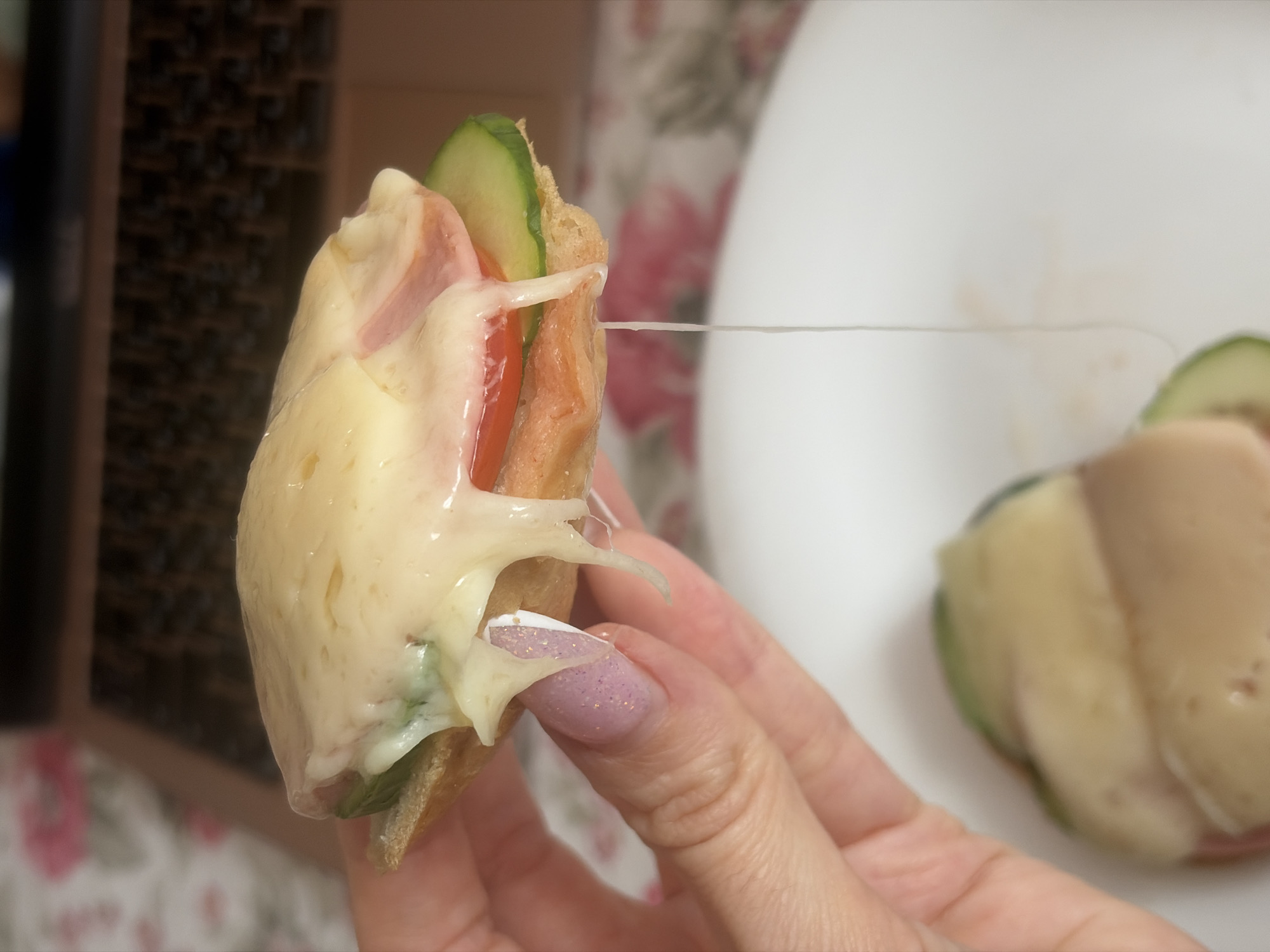 A close-up shot depicts a hand holding a small sandwich with melted cheese. The sandwich contains bread, melted cheese, sliced cucumbers, tomato slices, and a layer of meat. The cheese is stringy and drips down the sides of the sandwich. The hand has light skin and a decorated fingernail. Another similar sandwich is visible in the background, slightly out of focus, resting on a white surface. The background includes a dark, textured surface and a floral patterned surface. The city of Saint Petersburg, Russia, is not discernable in this image.