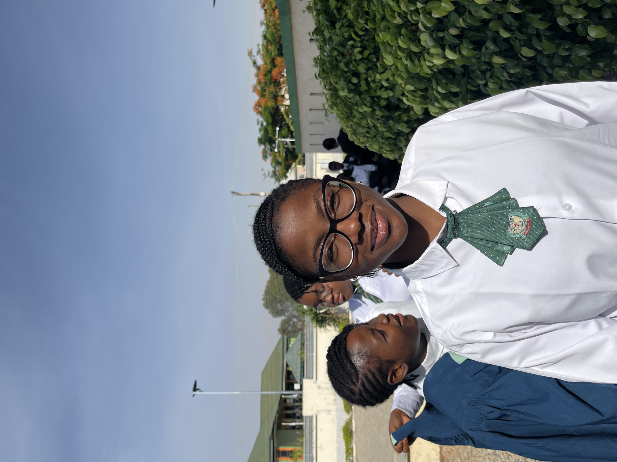 The image shows a close-up of four young people, likely students in Gura Top, Nigeria, wearing white shirts and green patterned ties. The foreground features a young woman with dark hair and glasses, smiling at the camera. Behind her is another young woman with braided hair whose eyes are closed and who appears to be resting on her friend's shoulder. A third person is partially visible behind them. In the background, there is a group of people wearing similar attire, and the setting appears to be outdoors, with green foliage and a light blue sky visible. A building with a green roof is also visible in the background.