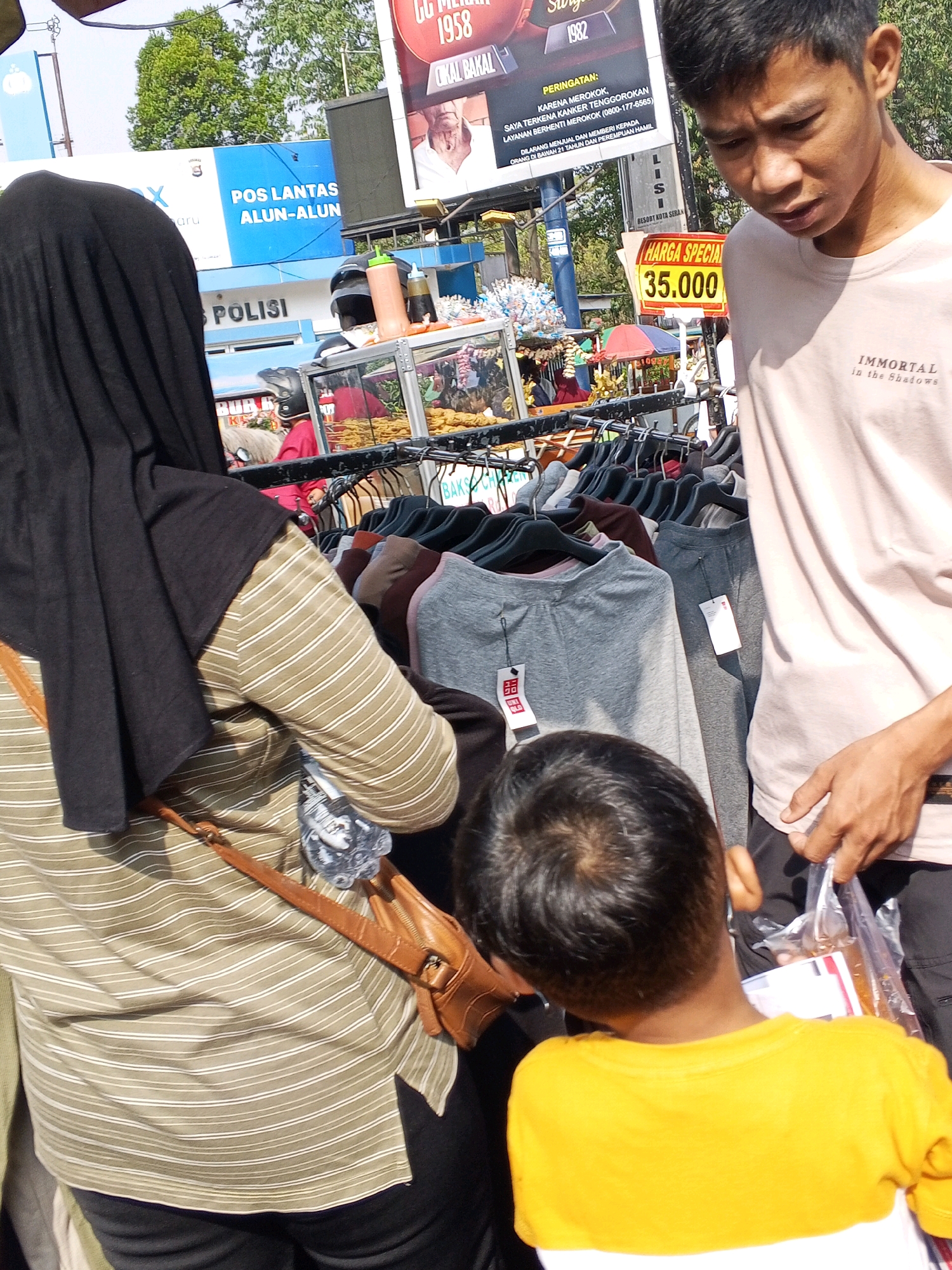 The image depicts an outdoor market scene, possibly in Serang, Indonesia, with a focus on a clothing stall. In the foreground, a person wearing a black hijab and a striped long-sleeved shirt is seen from the back, with a brown shoulder bag. To the right, a young man wearing a light pink t-shirt with "IMMORTAL in the Shadows" printed on it is interacting with another person whose back is to the camera. This second person is a child wearing a yellow t-shirt. In the background, a food stall with various items, including what appears to be fried snacks, is visible. Behind the food stall, a sign for "POS LANTAS ALUN-ALUN POLISI" is prominent, indicating a police traffic post located at the alun-alun (town square). A large billboard with text in Indonesian is also present. The overall environment suggests a bustling daytime market setting.
