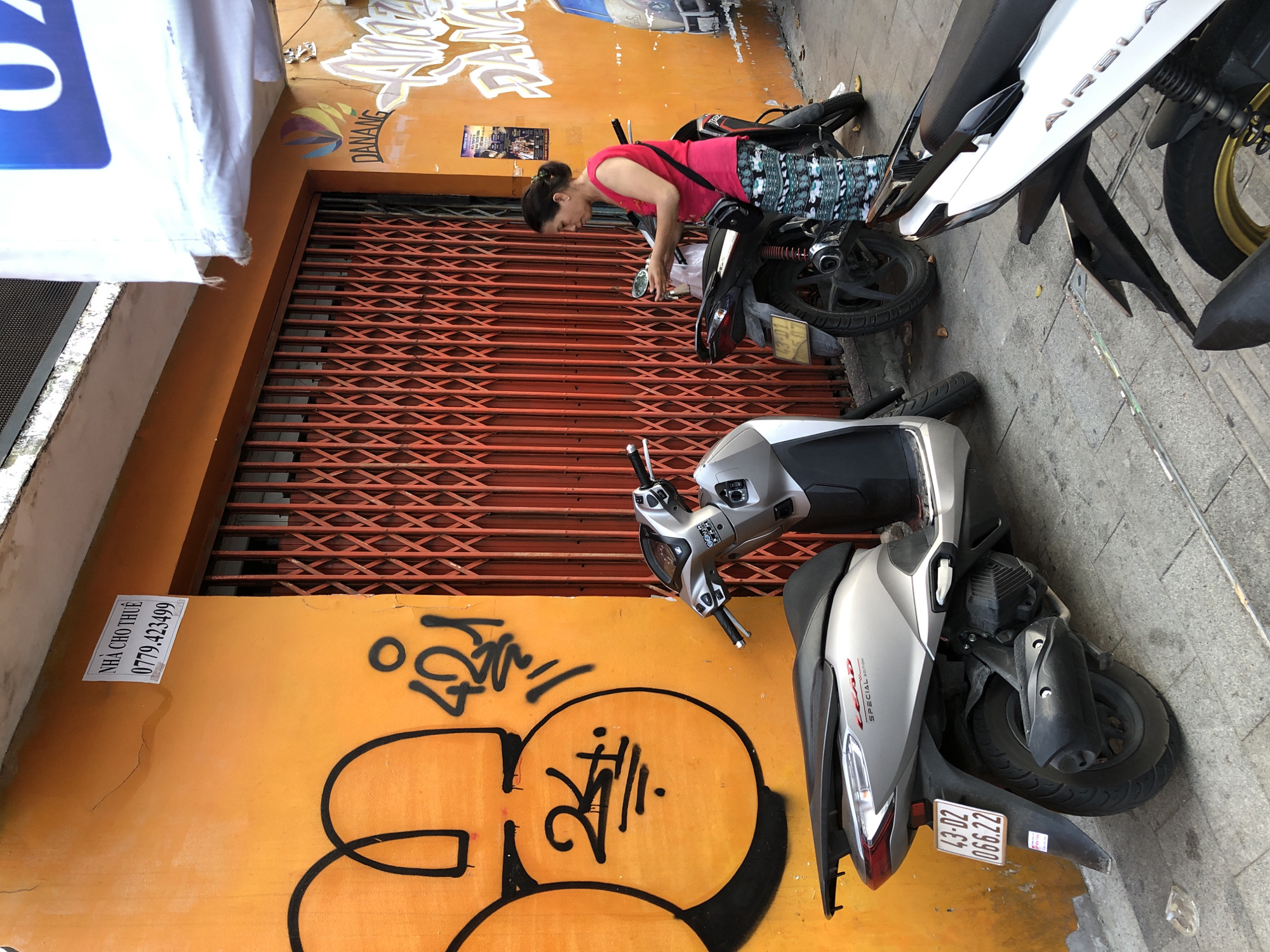 A woman in a pink sleeveless shirt and patterned pants stands beside a black motorcycle, holding a mobile phone. She is on a city sidewalk in front of a building with an orange wall covered in graffiti. Behind her, a silver scooter is parked. An orange metal security gate is closed. A sign on the orange wall reads "DANANG". Another sign in English states "NHA CHO THUE" and a phone number. The ground is paved with concrete.