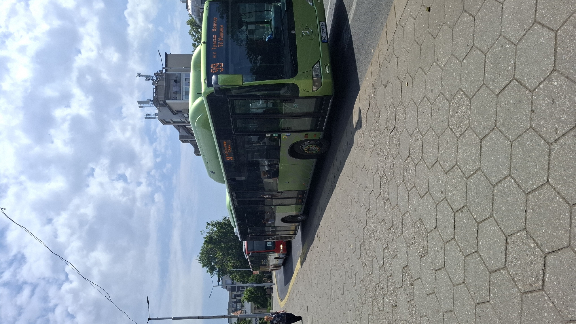 In Plovdiv, Bulgaria, a street scene unfolds on a bright, partly cloudy day, with a person walking on a hexagonal-tiled sidewalk in the foreground. Behind them, a line of buses is visible. The foremost bus is a bright green Mercedes-Benz articulated bus, displaying the route number "99" and destinations "Ж.К. Тракия - Център" and "ТК Мариса". Its license plate reads "PB 1299 TE". Further down the road, a red bus with the route number "37" is also present. In the background, buildings with telecommunication antennas on their roofs, trees, and a street light contribute to the urban environment. The sky is a mix of blue and white clouds.