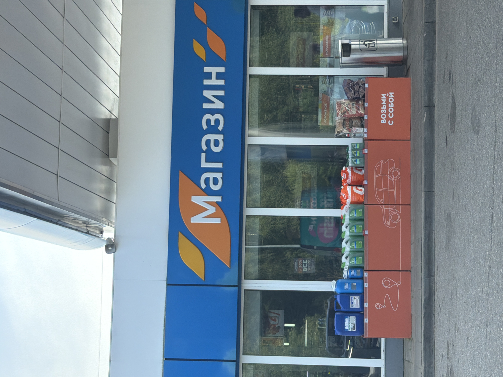 The image shows the facade of a store with a large blue sign that says "МАГАЗИН" (MAGAZIN) in white letters and orange graphic elements. Below the sign are large windows displaying various products. In front of the windows, there is a red display unit with a white outline of a car and a path with location pins. On top of this red unit, bottles of various fluids, bags of charcoal, and other items are displayed. To the right of the display unit is a silver trash can. The store appears to be a convenience store or a shop at a gas station. The overall environment suggests an outdoor setting, likely in a developed area. The image is taken from a slightly low angle, looking up towards the store. The lighting suggests it is daytime. There are no visible people in the image.