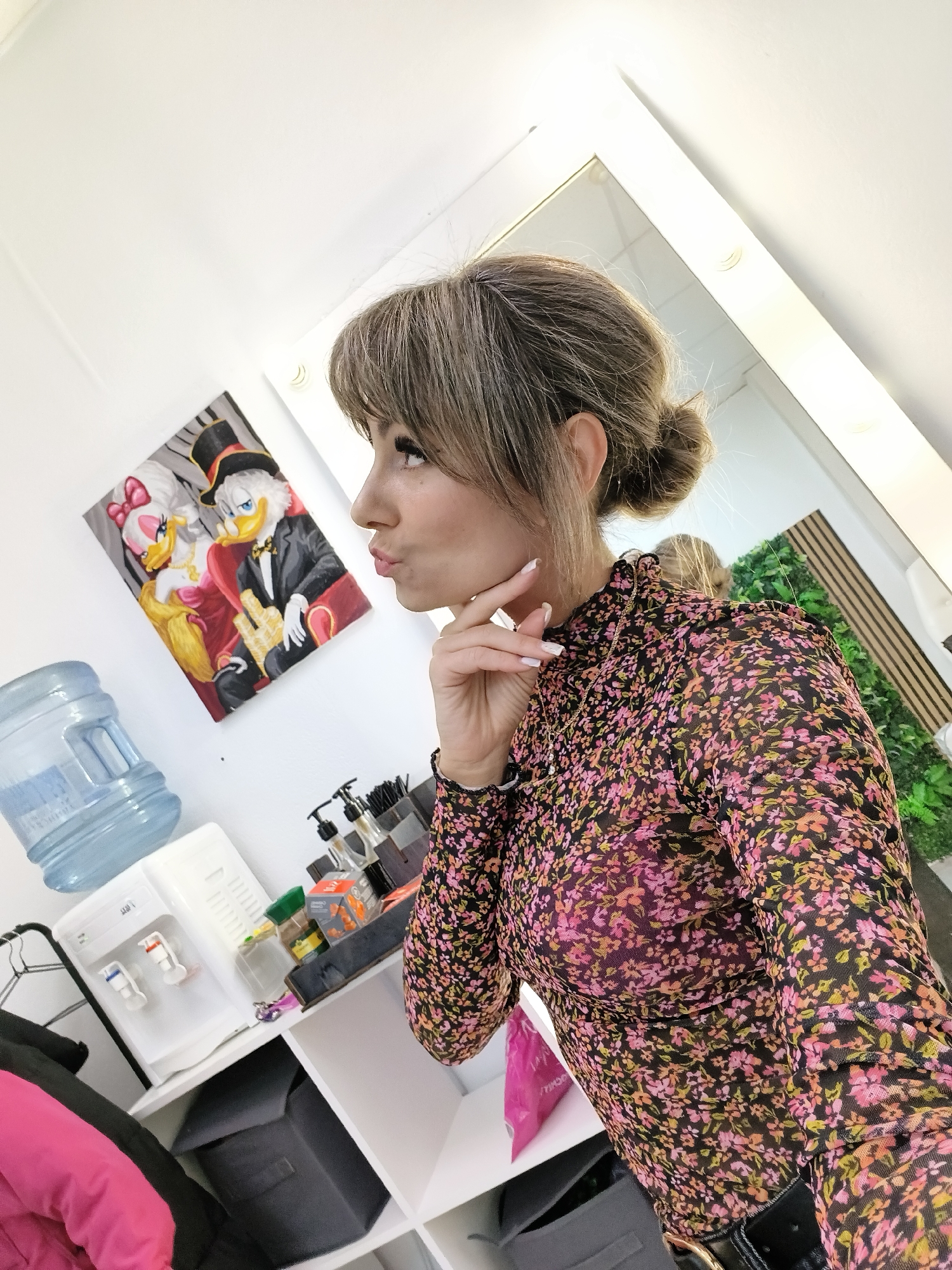 A woman with light brown hair, styled in a bun with bangs, is the primary subject of this indoor photo. She is wearing a high-necked, long-sleeved top with a black background and a dense floral pattern in shades of pink, orange, and yellow. A delicate gold chain necklace is visible, and her fingernails are painted white. She is captured in a playful pose, looking upwards to her right with pursed lips in a "duck face" expression, her left hand gently touching her chin.
The setting appears to be a brightly lit room, possibly a dressing room or studio. On the white wall behind her, to the left, a painting depicts two anthropomorphic ducks resembling Disney characters: a male duck in a tuxedo and top hat, and a female duck in a red dress with a large bow. Below the painting, a large blue water bottle rests on a white water cooler/dispenser, which features red and blue spigots and the visible text "АЙС." A white shelving unit next to it holds several bottles with pump dispensers and a dark fabric storage bin. A pink jacket and other clothing items are partially visible in the bottom left corner.
To the woman's right, a large mirror with illuminated bulbs along its white frame (a vanity mirror) reflects a wall covered in artificial green foliage. The scene conveys a candid moment in a functional, well-lit space in Chaikovsky, Russia.