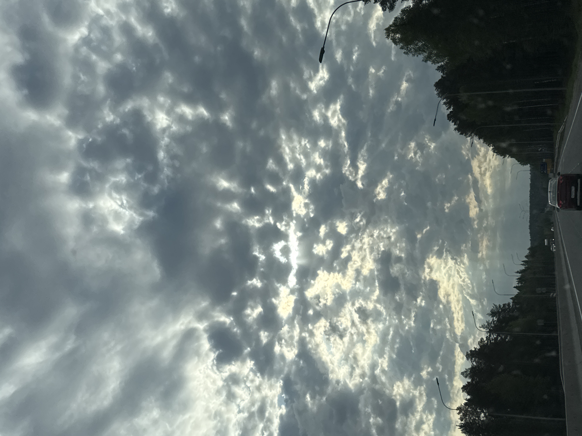 The image depicts a view from a vehicle traveling on a road. The sky is predominantly filled with a dramatic display of altocumulus clouds, through which sunlight is breaking. The road has streetlights, and there is a line of trees bordering it. In the distance, other vehicles can be seen on the road. The location could potentially be in or near Белобородово, Russia, based on the information provided.