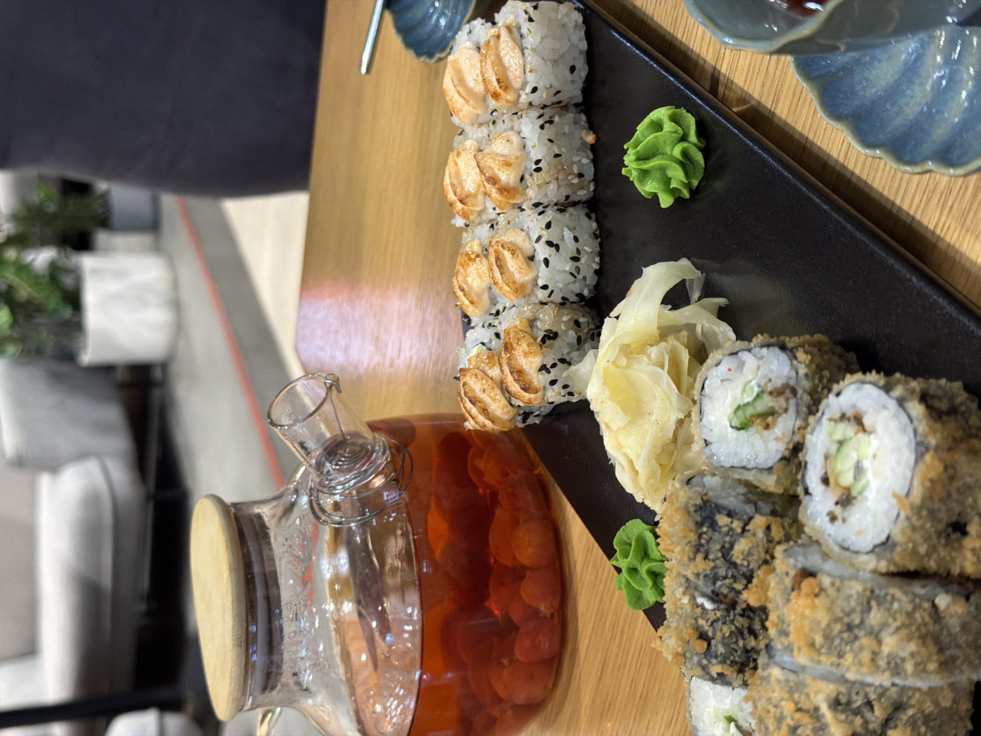 A glass teapot with a wooden lid filled with a red beverage and fruit sits on a wooden table next to a black rectangular plate of sushi. The sushi is arranged in two rows. The top row consists of several pieces topped with seared salmon. The bottom row includes fried sushi rolls with visible cucumber and cream cheese inside. Accompaniments to the sushi include pickled ginger and a dollop of bright green wasabi. In the background, blurred furniture and plants suggest an indoor setting, possibly a restaurant.