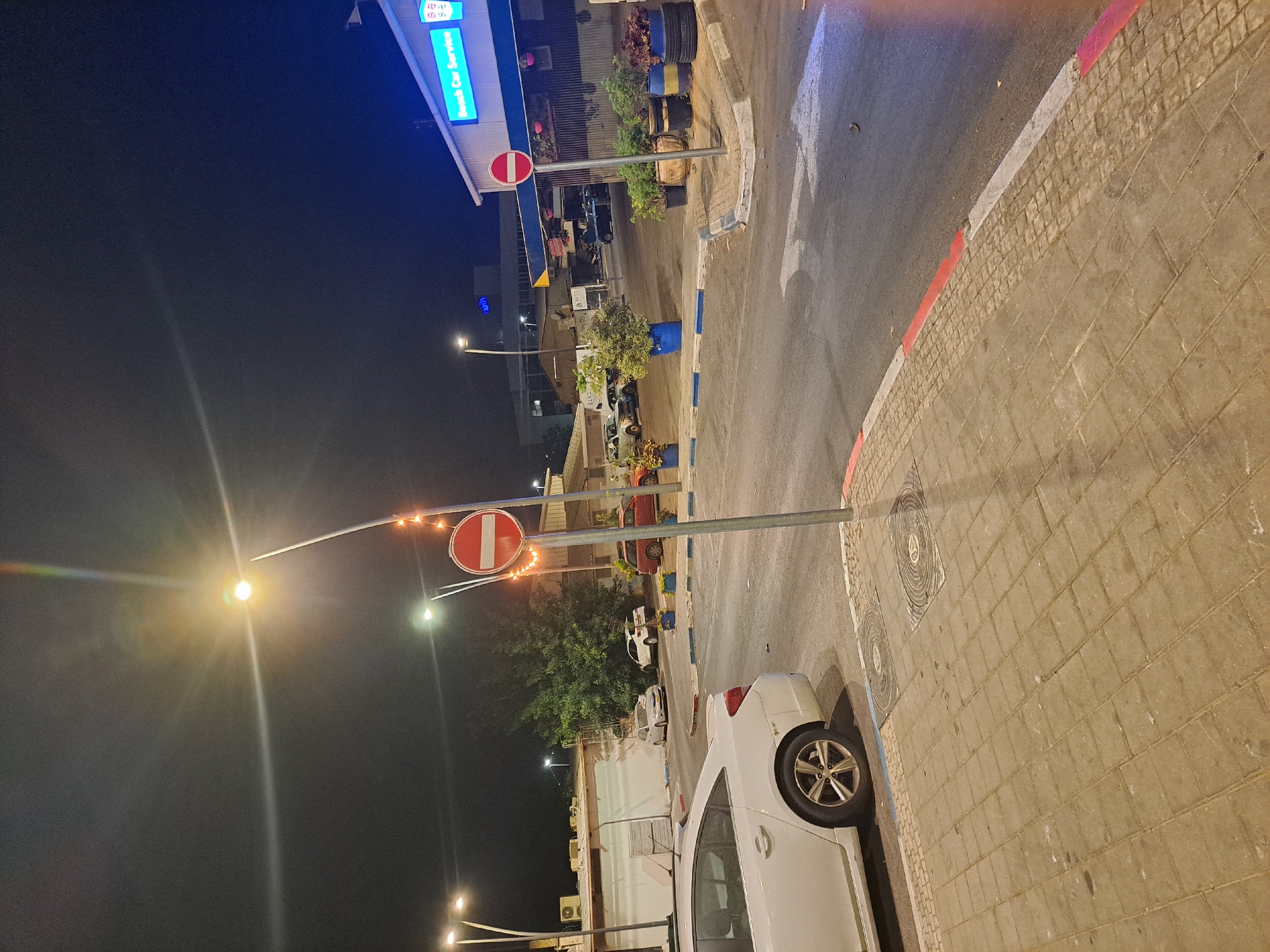 The image depicts a nighttime scene in the city of Ramat HaSharon, Israel, specifically at a location that appears to be a street or intersection near a car service establishment.
In the foreground, a white car is parked to the left, facing away from the viewer. The car's rear wheel and a portion of its side are visible. The ground in the foreground is paved with rectangular interlocking stones, with manhole covers embedded within. A red and white striped curb runs along the edge of the road.
The road itself is asphalt, with white lane markings and arrows visible. To the right of the road, a street lamp illuminates the area. A prominent circular traffic sign, red with a white horizontal bar, is positioned on a pole adorned with small orange string lights. Several large potted plants are arranged along the sidewalk area.
In the middle ground, several other cars are parked or in transit. A building with a blue and white sign reading "Bosch Car Service" is visible to the right, indicating a business. Another building with an air conditioning unit is present to the left. Trees and foliage are scattered throughout the scene.
The background shows more buildings and streetlights, suggesting an urban environment. The sky is dark, characteristic of nighttime. The overall impression is of a well-lit street in a commercial or residential area at night.