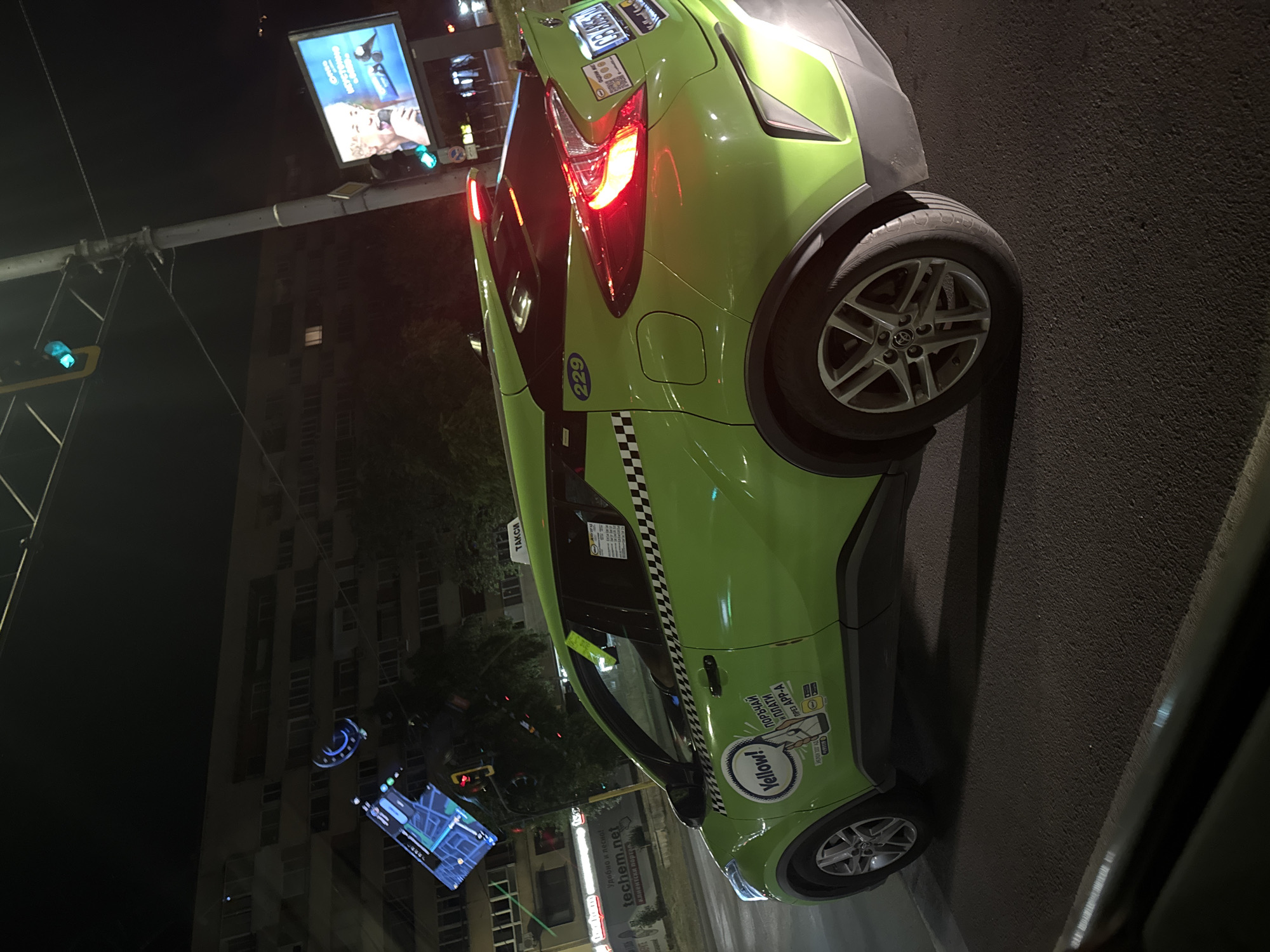An urban scene captured at night in Sofia, Bulgaria, shows a lime green taxi, a Toyota C-HR, with the "Yellow!" logo prominently displayed on its side. The taxi has a checkerboard stripe along its body and a "TAKSI" sign on its roof. It is parked on an asphalt road, with buildings in the background illuminated by streetlights. To the right of the taxi, a large digital billboard displays an advertisement for a person holding a microphone. Traffic lights, one showing green, are visible above and to the left of the billboard. A navigation screen is mounted on the taxi's dashboard and is visible through the windshield, displaying a map. The image also shows a traffic light hanging from a pole, and wires crisscrossing the sky.