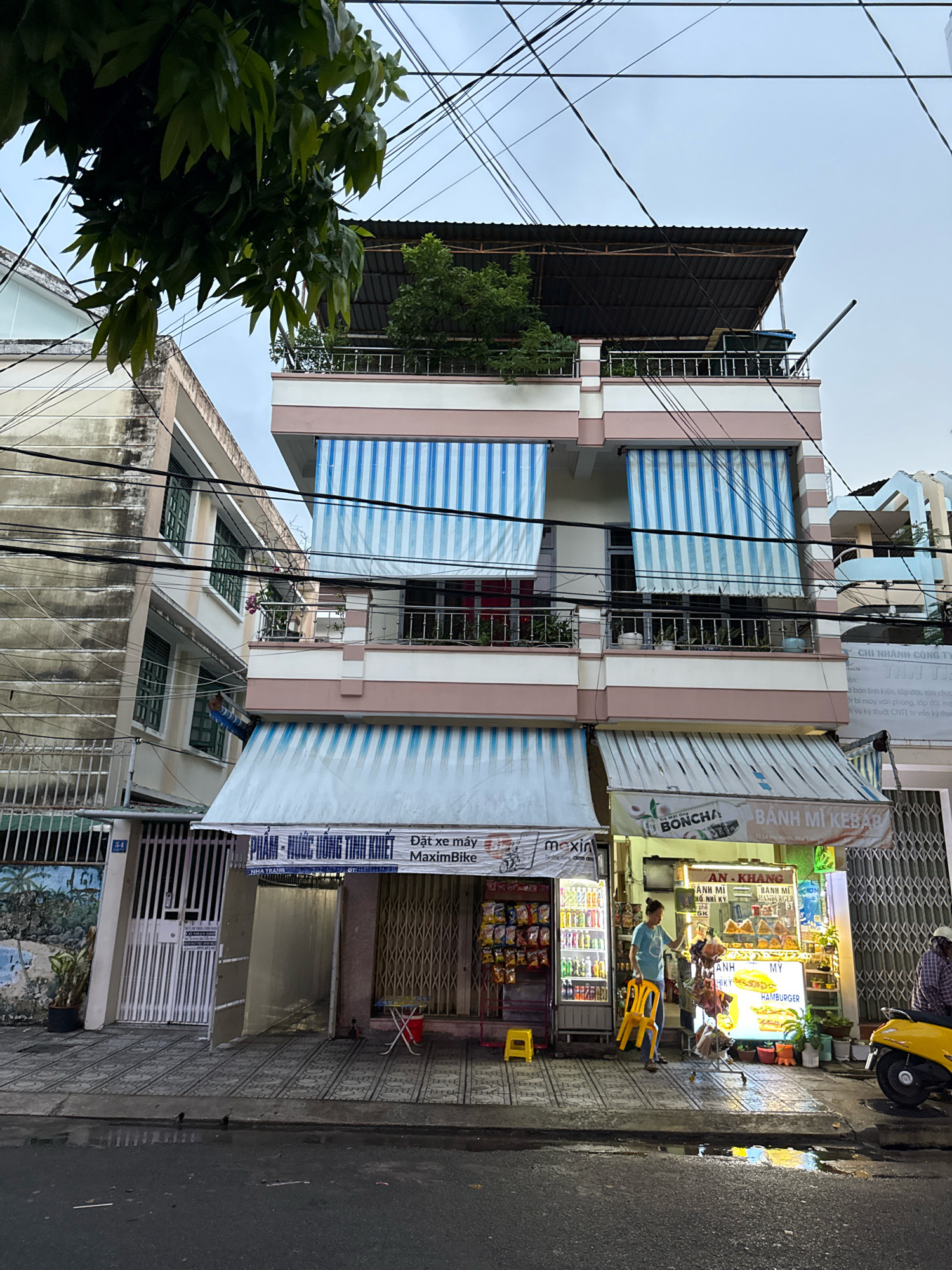 The image shows a street scene in Nha Trang, Vietnam, featuring a multi-story building with shops at the ground level and residential units or balconies above. The building has a light pink and white facade with blue and white striped awnings covering the shopfronts and balconies.
On the ground floor, there are at least two businesses visible. To the left, a shop with a white gate and a mural depicting palm trees is present. Next to it, a food stall is brightly lit, displaying various food items and signage in Vietnamese. This food stall appears to be selling bánh mì kebab, hamburgers, and other Vietnamese dishes. A woman in a blue t-shirt and dark pants is standing in front of the food stall, holding a yellow plastic chair. A yellow scooter is parked to the right of the food stall, and a person wearing a checkered shirt is seated on it.
The street in front of the shops is paved with decorative tiles, and there are some wet patches on the ground, suggesting recent rain. Power lines are visible crisscrossing above the buildings. Trees with green foliage are present in the foreground and on a rooftop terrace. The sky is overcast, indicating a cloudy or potentially damp day. The architecture is typical of urban areas in Vietnam, with narrow buildings and a mix of commercial and residential spaces.