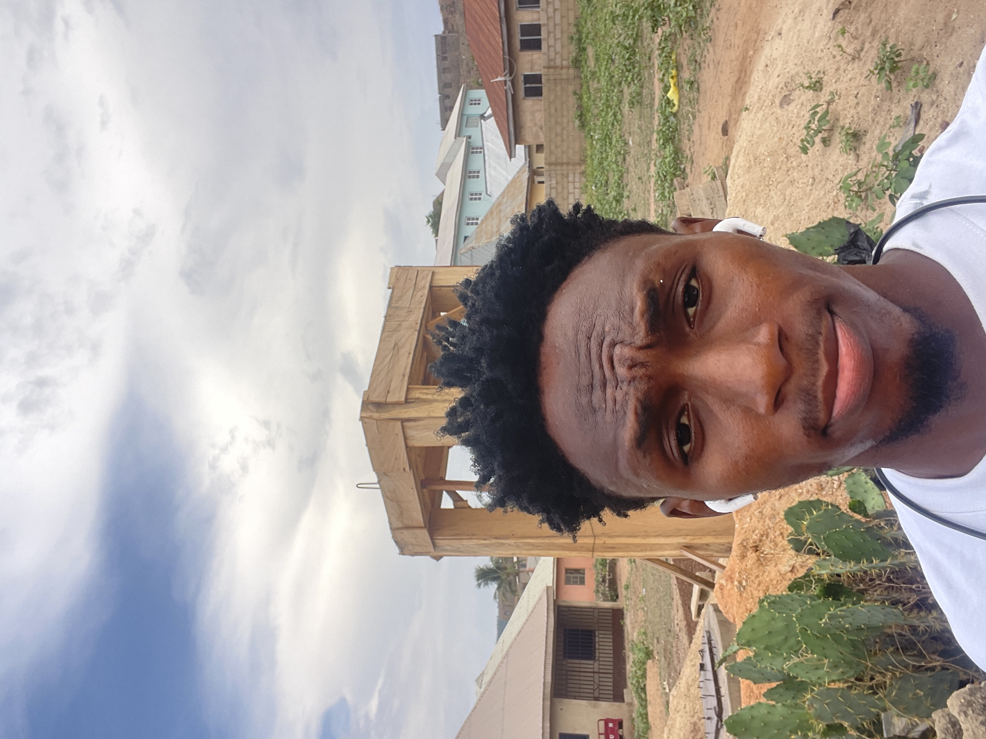 A young Black man is captured in a selfie-style portrait, looking directly at the viewer with a slightly intense or pensive expression, his brow furrowed. He has short, dark, curly hair and a subtle beard/stubble. He is wearing a white t-shirt with a dark trim and white wireless earbuds in both ears.
The background reveals an outdoor, semi-urban or rural setting under a partly cloudy sky with patches of blue. Behind him, a wooden scaffolding-like structure is visible, suggesting ongoing construction or a utility stand. Beyond this, several buildings with corrugated metal roofs and concrete block walls are scattered across the landscape. Some structures appear to be residential, with windows and light blue siding on one building. The terrain consists of bare earth or sandy patches mixed with green vegetation, including a prominent cactus plant with spines in the immediate foreground to the man's right. The overall impression is a daytime scene in a developing area, consistent with Lokoja, Nigeria. There is no visible text in the image.