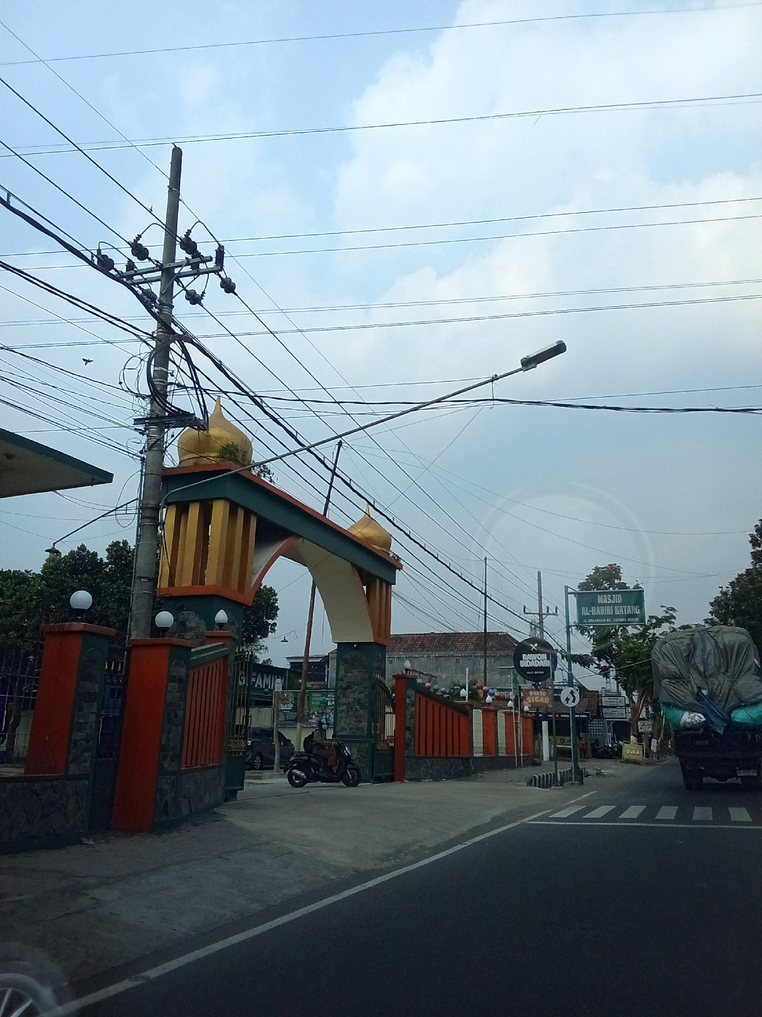 The image shows an archway with two golden domes, likely the entrance to a mosque, in Ngasem, Indonesia. The archway is decorated with orange and green accents. Behind the archway, a sign reads "MASJID AL-HABIBI KATANG". A person is riding a motorcycle through the entrance. The scene is characterized by a cloudy sky, numerous electrical wires, a utility pole, and trees. To the right, a truck covered with a tarp is parked on the side of the road. A pedestrian crossing is visible in the foreground. Other signs for businesses, such as "RAWON BIDADARI" and "BAKSO SANGAR", are also present. The overall environment appears to be a street in a semi-urban area.