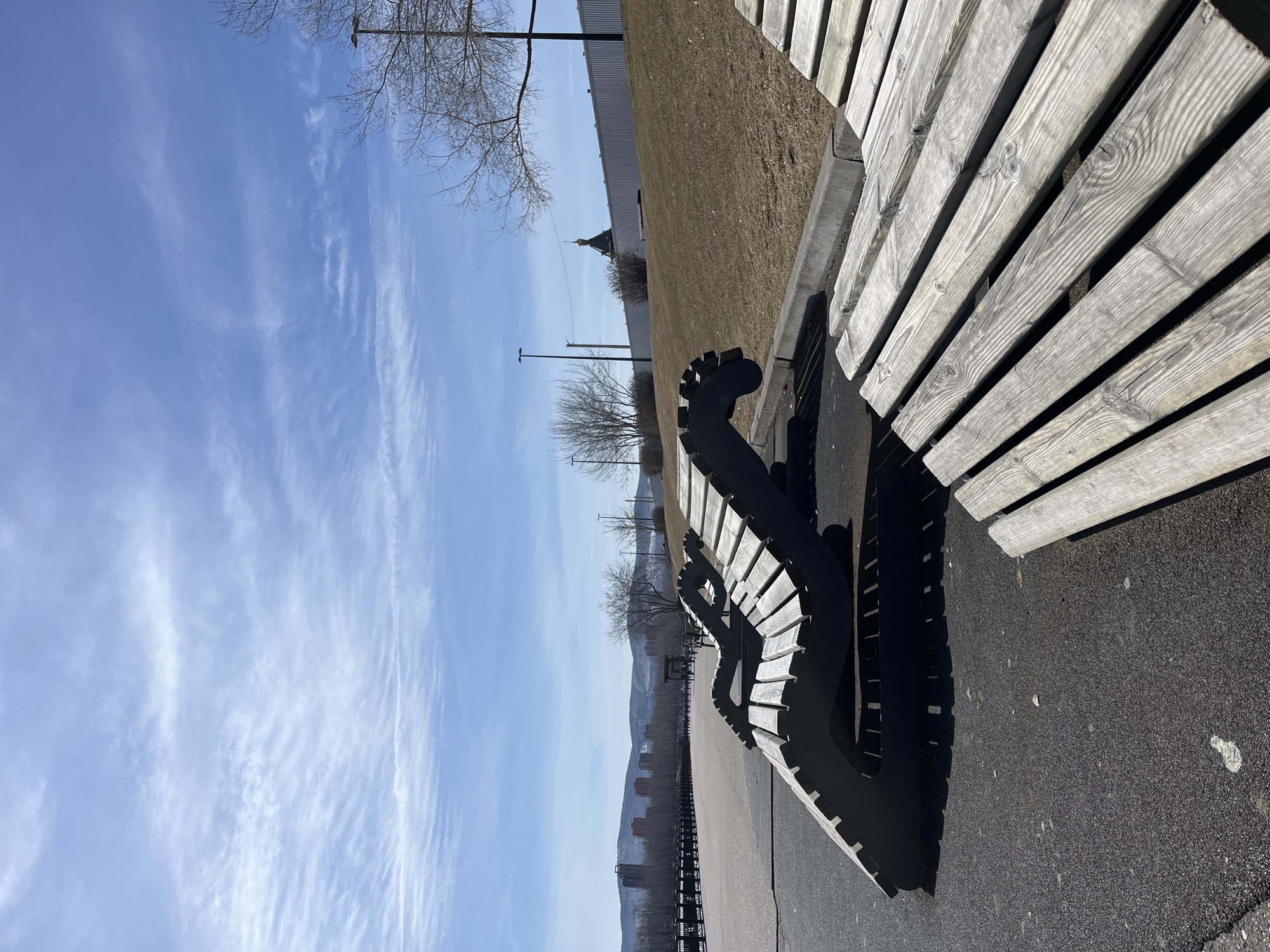 The image depicts a row of benches at a park in the city of Krasnoyarsk, Russia. The benches are made of wooden planks and have a unique, curvilinear black metal frame. The park appears to be located along a waterfront or riverbank, with a low fence visible in the background. Beyond the park, the city skyline is faintly visible in the distance with buildings and possibly hills or mountains. A building with a distinctive cupola is visible along with utility poles and bare trees. The sky is blue with wispy clouds. The ground is paved with asphalt or a similar material.