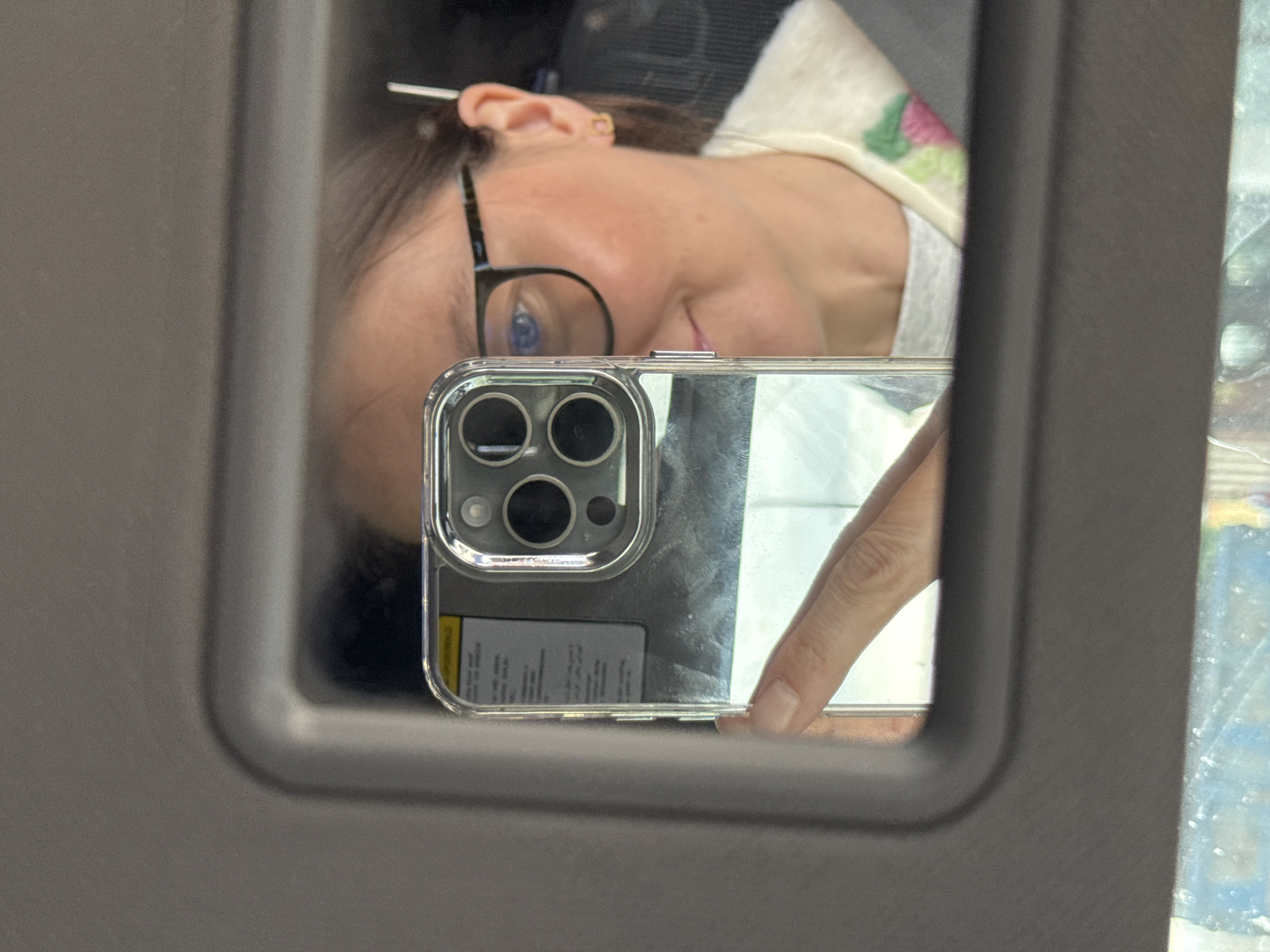 The image captures a mirror selfie taken from inside a vehicle, likely a car. The primary subject is a person, reflected upside down in the mirror, which is framed by dark gray plastic, typical of a car's sun visor or rearview mirror.
The person has dark hair, pulled back and secured with a white clip or stick. They are wearing black-rimmed glasses, which highlight their blue eyes. A subtle, neutral expression is visible on their face. Part of their upper body is visible, showing a light-colored garment, possibly a t-shirt or sweater, with a faint floral or patterned design featuring pink and green elements on the shoulder.
A hand with natural-colored nails is holding a smartphone, which has a distinctive triple-camera setup on its back, suggesting it's an iPhone, enclosed in a clear protective case. The phone is positioned to capture the mirror reflection. On the back of the phone, beneath the camera module, a yellow sticker with blurred, unreadable text is visible through the clear case.
The background, seen through the mirror, is a blurry outdoor scene with natural light, indicating it is daytime. The overall setting is an interior of a vehicle, consistent with the location context of Izhevsk, Russia. The activity is a casual mirror selfie.