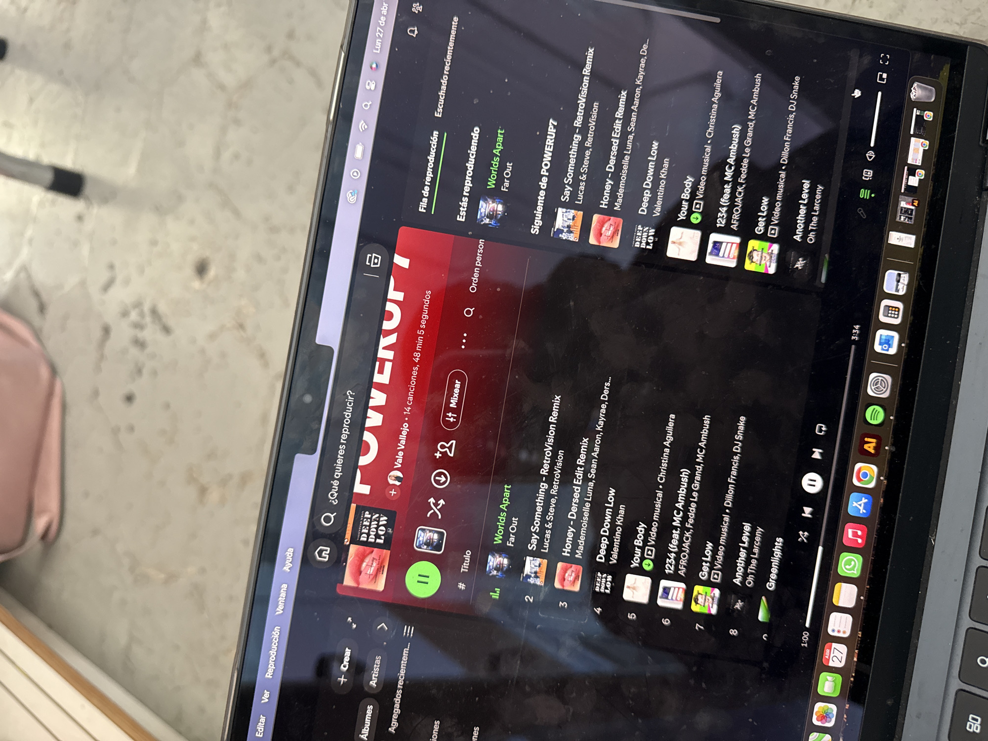 The image displays a laptop screen showing the Spotify music application, set against a light-colored, possibly tiled, surface. The screen is visibly smudged and dirty. The application interface is in Spanish, with menu options like "Editar," "Ver," "Reproducción," "Ventana," and "Ayuda."
The "POWERUP7" playlist, created by Vale Vallejo, is active, comprising 14 songs with a total duration of 48 minutes and 5 seconds. The song "Worlds Apart" by Far Out is currently playing, with playback paused. The player's progress bar indicates a timestamp of 1:00.
The "Siguiente de POWERUP7" (Next in POWERUP7) queue lists several tracks: "Say Something - RetroVision Remix" by Lucas & Steve, "Honey - Dersed Edit Remix" by Mademoiselle Luna and others, "Deep Down Low" by Valentino Khan, "Your Body" by Christina Aguilera (noted as a music video), "1234 (feat. MC Ambush)" by AFROJACK, Fedde Le Grand, and MC Ambush, "Get Low" by Dillon Francis and DJ Snake (also a music video), "Another Level" by Oh The Larceny, and "Greenlights".
The laptop's system bar shows the date "Lun 27 de abr" (Monday, April 27th) and the time 3:34, likely indicating 3:34 PM. Various application icons, including Spotify and WhatsApp, are visible in the dock at the bottom right. A blurry pink object is partially visible on the left side of the frame.