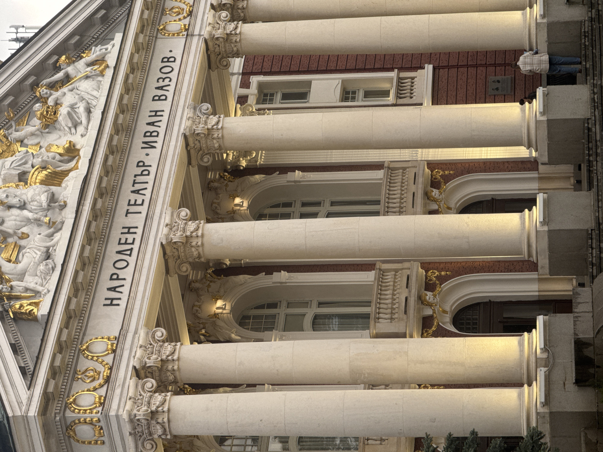 The image shows the National Theatre Ivan Vazov in Sofia, Bulgaria. The photo captures the façade of the building, featuring a row of classical columns. The building is adorned with golden sculptures and ornaments. The inscription "НАРОДЕН ТЕАТЪР · ИВАН ВАЗОВ" is visible. Two people stand on the building's balcony. The exterior architecture is illuminated with subtle lighting.