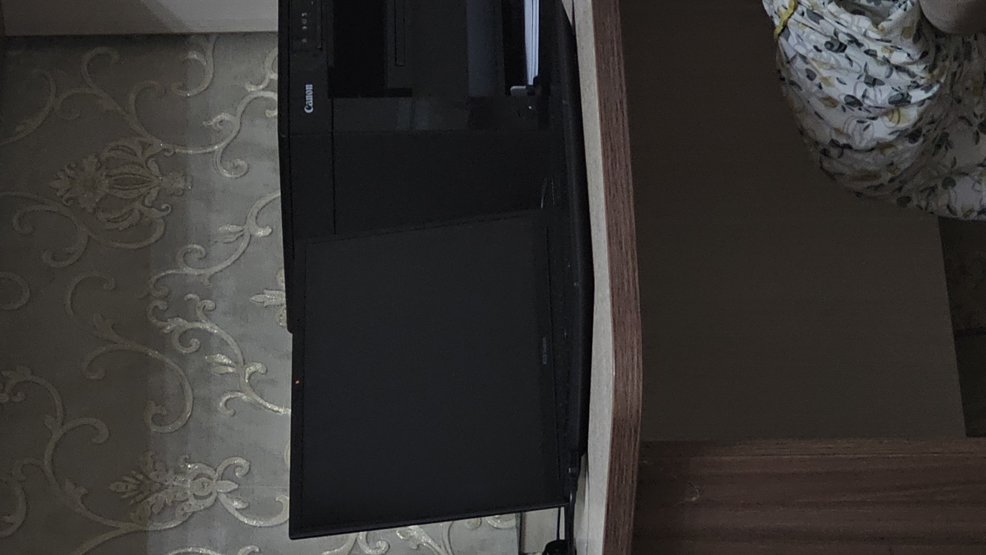 In Yandare, Russia, a black laptop is positioned on a wooden desk against a wall with ornate wallpaper. The laptop is open, displaying a dark screen, and appears to be powered on, indicated by a small red light visible on the bezel. To the right of the laptop, a black Canon printer is present. The environment suggests an indoor setting, possibly a home office or study. The lighting is dim, with the primary light source appearing to be ambient room light.