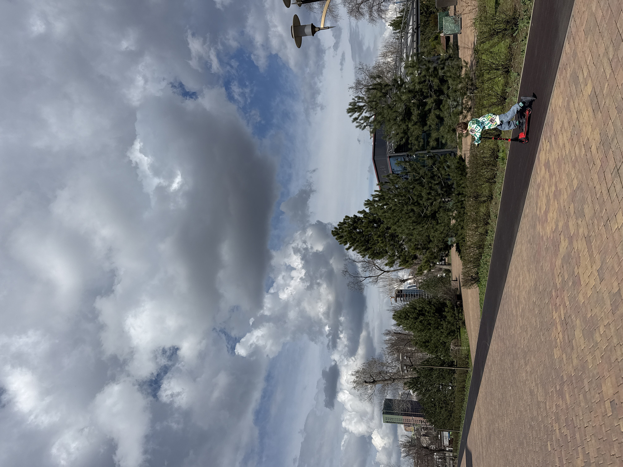 A sideways, medium outdoor shot shows a young person on a red scooter on a bricked path, near a planted green space, set against an overcast sky, likely taken in Rostov-on-Don, Russia.
The main subject, a child or young teenager, is seen riding a red scooter. They are wearing jeans and a white patterned jacket. The angle of the shot suggests they are moving from the upper left to the lower right of the frame.
The environment includes a bricked pathway which has a dark border. There is a green strip planted with trees and other plants alongside the path. Buildings, some high-rise, are seen in the background, and street lamps are visible near the green space.
The sky is filled with gray and white clouds, indicating an overcast weather condition. The setting suggests a park or pedestrian area within a city, and the architectural style and landscape point towards a location within Rostov-on-Don, Russia.
