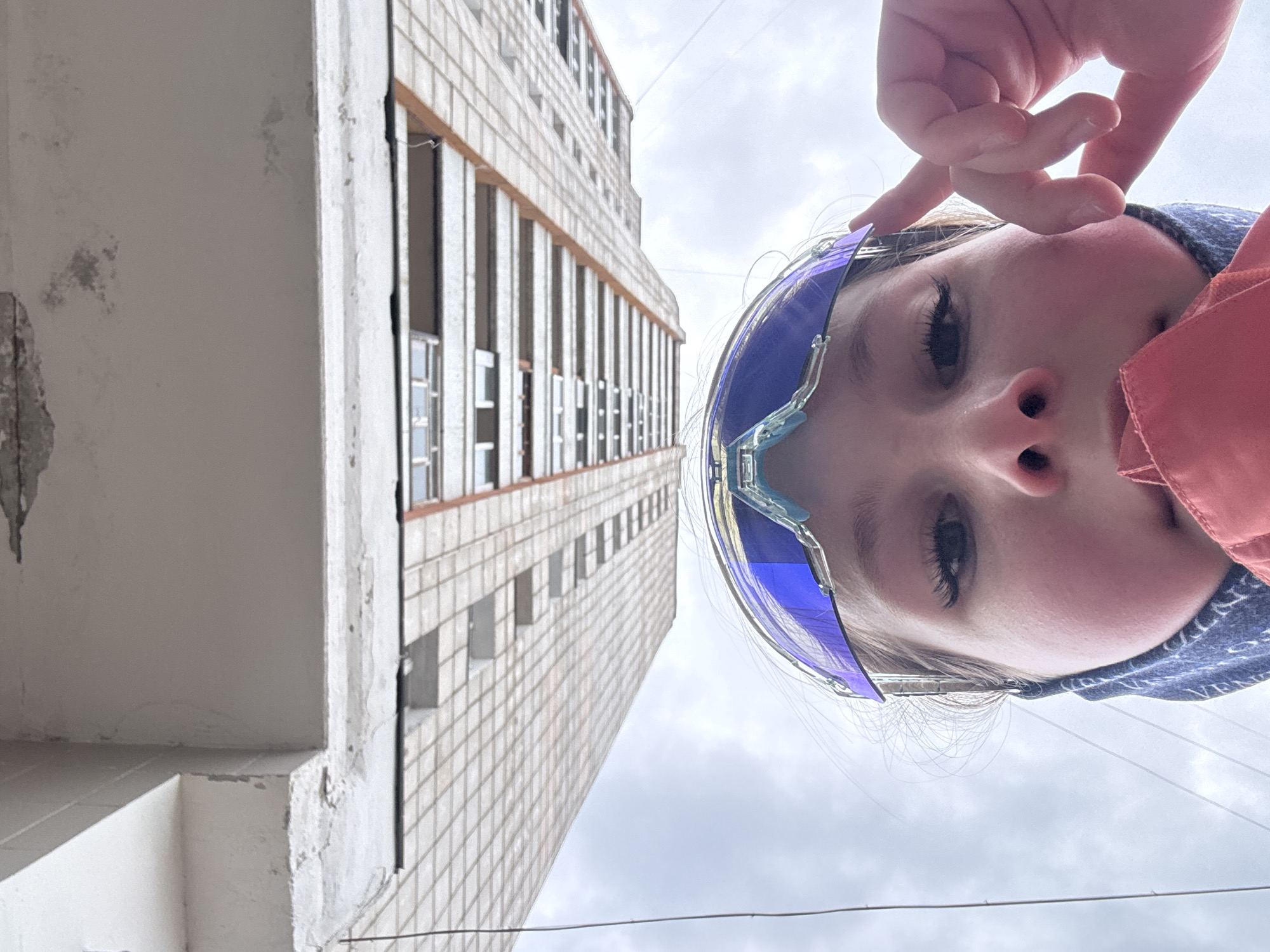 A low-angle shot of a child wearing blue protective eyewear and standing outside a building. The child's face dominates the foreground. A tall building with a grid-like facade, characteristic of Soviet-era architecture, rises behind and to the left. The sky is overcast. The building has multiple rows of windows and a worn, aged exterior, possibly tiled. Based on the architectural style, it is likely a residential or institutional structure. The location is Chaikovsky, Russia. There are electrical wires visible across the sky in the bottom portion of the picture.