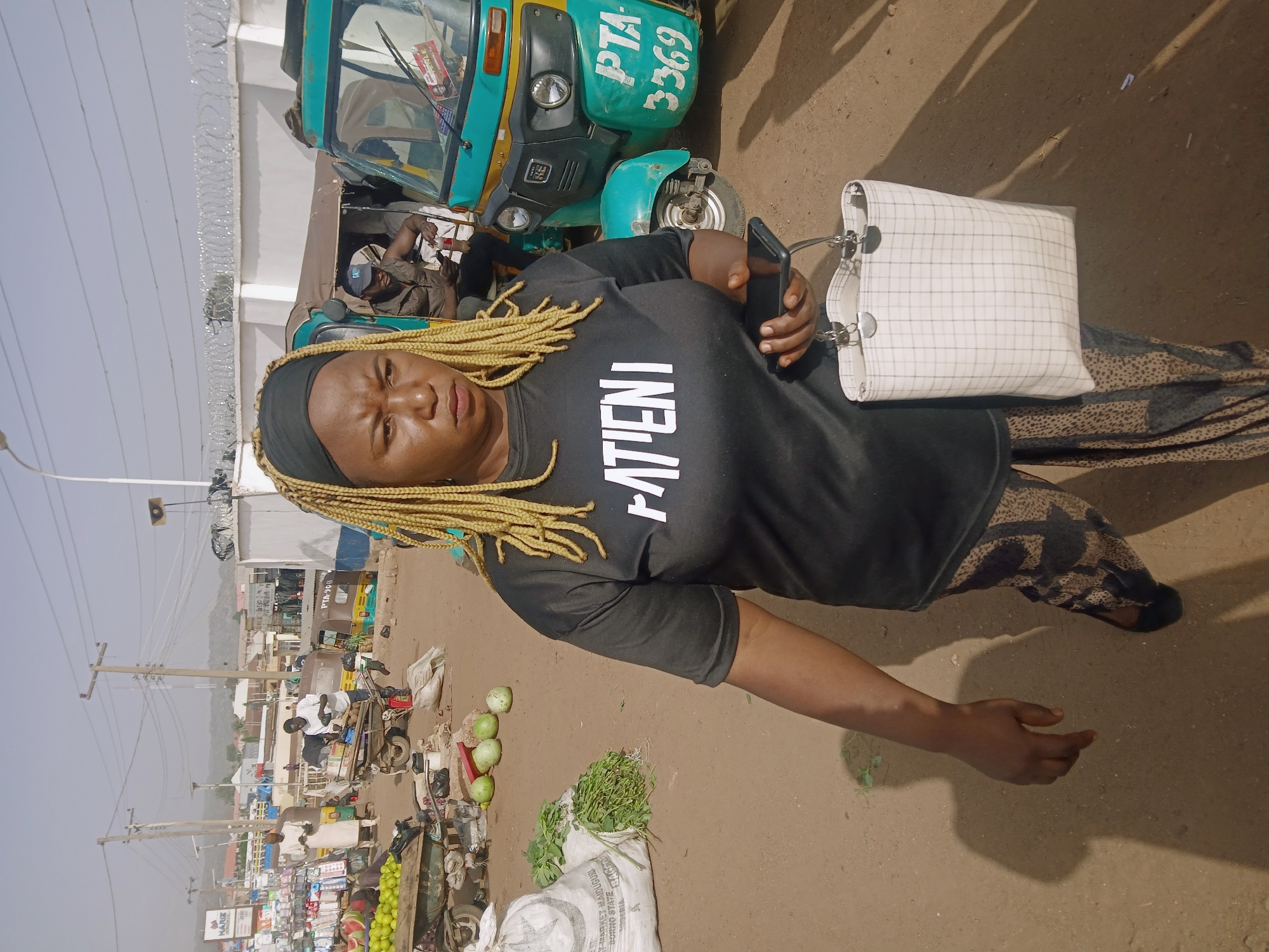This portrait-oriented image captures a woman in the foreground against an outdoor market scene in Rafin Par, Nigeria. The woman has light blonde dreadlocks or braids, a dark headwrap, and wears a black t-shirt with "PATIENT" printed vertically in white capital letters. She is also wearing patterned dark trousers and black shoes. She holds a black smartphone in her left hand and a white, grid-patterned handbag with metallic chain details in her right, looking directly at the camera with a neutral expression.
Behind her, the dusty, unpaved ground stretches into a lively market area. A green and yellow motorized trishaw, commonly known as a "keke napep," is prominently visible, bearing the license plate "PTA 3369". Another trishaw further back shows "PTA 864Y4". A man is seated inside the closest trishaw. Along the left side of the frame, market stalls display various produce, including green fruits, watermelons, and leafy vegetables, some packaged in sacks. Several people are visible in the background, including vendors and customers. Utility poles with numerous power lines crisscross the sky, which appears bright and clear, indicating daytime. Shadows on the ground suggest midday or early afternoon. A building with razor wire on its top edge is also visible. The scene depicts a bustling, everyday market environment.