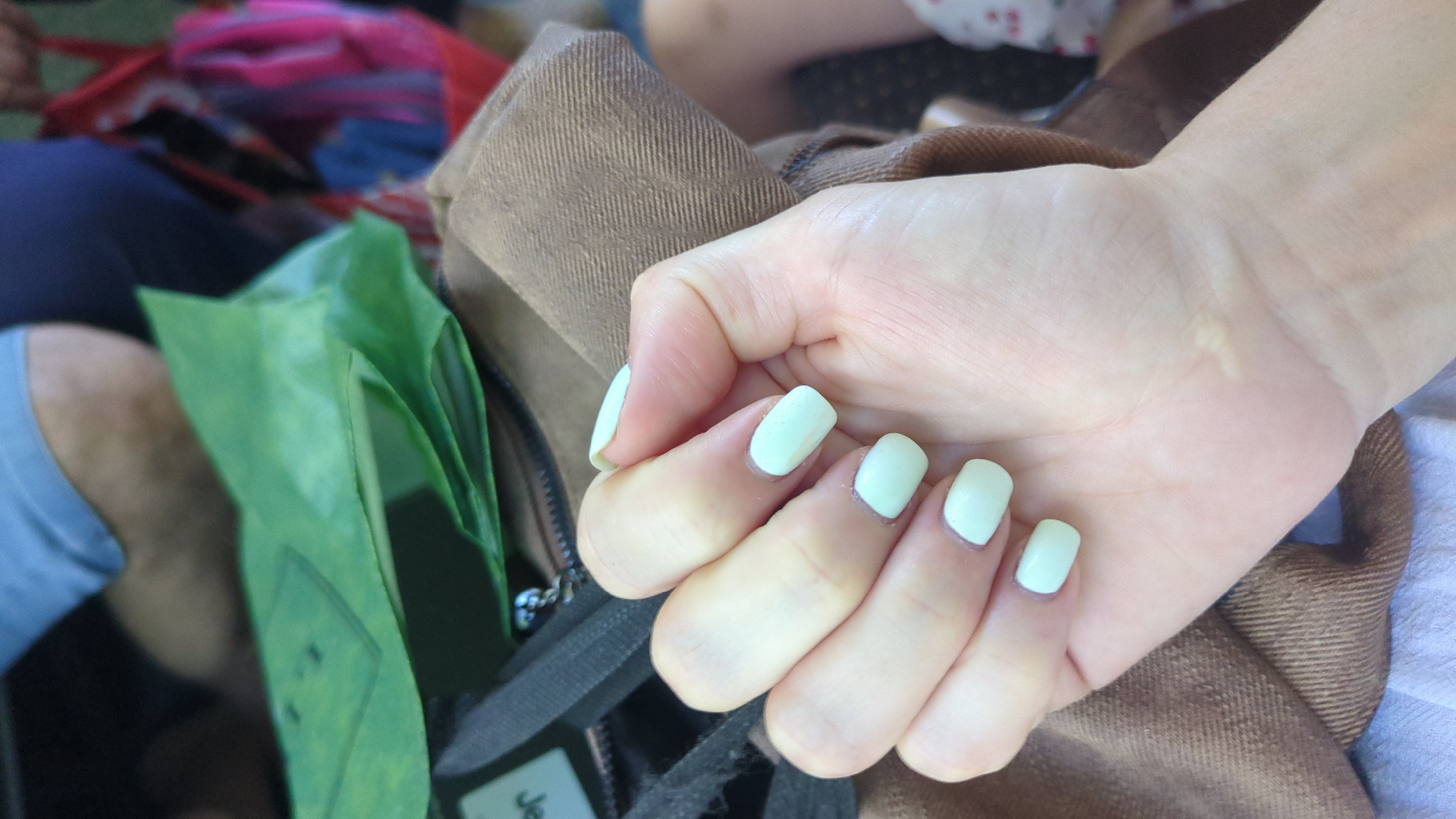 The image shows a close-up of a person's left hand with light-colored fingernails. The hand is resting on a brown fabric surface, likely part of a bag or clothing. In the background, partially visible, are other objects including a green plastic bag and glimpses of people in casual attire, suggesting an outdoor or public setting. The environment appears to be a city, possibly Chișinău, Moldova, indicated by the context provided in the prompt, though no specific landmarks are visible. The focus is on the hand and its manicured nails, with other elements providing context.