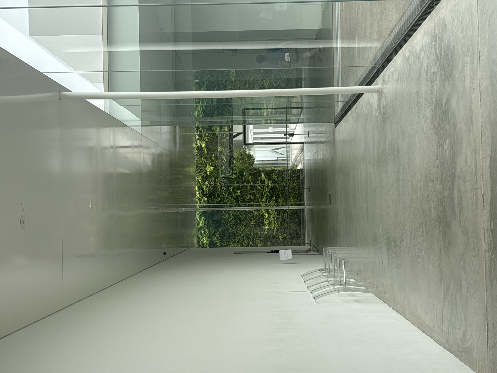 The image displays a bright, modern interior space, likely a gallery or public area within a building in Kanazawa, Japan. The perspective looks down a long, well-lit corridor or room.
On the left, a clean, smooth white wall extends into the distance, reflecting light. A horizontal white pipe or railing is attached to this wall. The ceiling above is also white and highly reflective.
On the right, a light grey concrete-textured wall provides a contrasting, somewhat industrial aesthetic. Along this wall, a row of approximately six transparent, possibly acrylic, chairs with curved backs is neatly lined up, suggesting a waiting or viewing area.
The central part of the image features expansive glass panels, acting as a window or partition. Through these panels, a lush, vibrant green living wall (vertical garden) is prominently visible, adding a natural element to the modern design. The glass surfaces exhibit significant reflections, showing distorted views of the interior white walls, parts of the concrete wall, and faint reflections of people within the building. One reflection in the upper glass shows a person or persons in dark attire, possibly a child or someone bending over. Another reflection of an upper body is visible closer to the green wall.
On the white floor, near the base of the living wall, a small, white rectangular sign or information stand is placed. Any text on it is too small and blurry to be clearly legible, though faint black markings suggest writing, possibly "EXIT" or directional information. The lighting indicates it is daytime. The overall scene conveys a sense of minimalist design, spaciousness, and serene modernity.