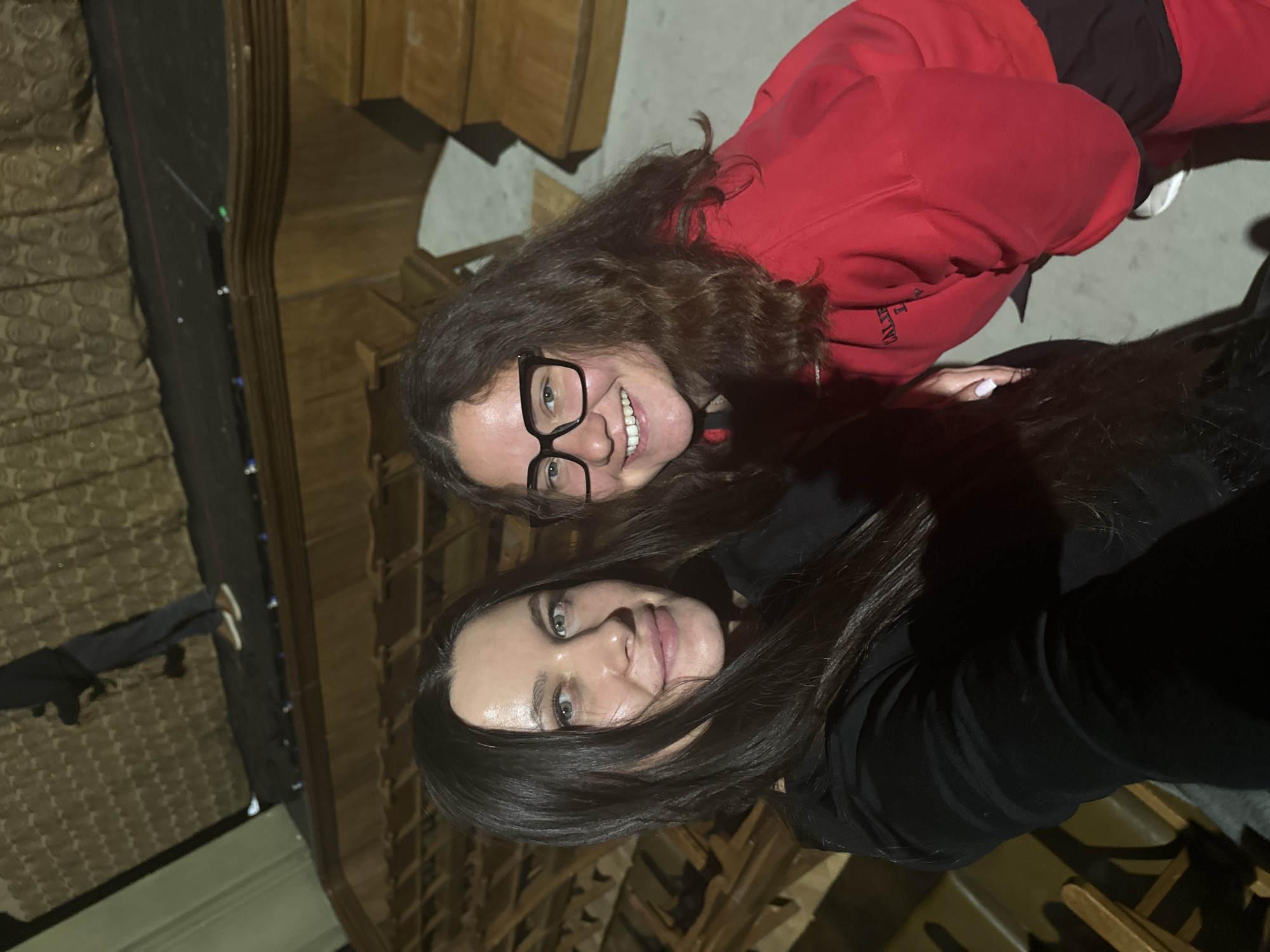 Two women pose for a selfie inside the Bolshoi Theatre in Moscow, Russia. One woman is wearing a red sweatshirt and glasses, the other a black jacket. They are both smiling. The seating of the theatre is visible in the background.