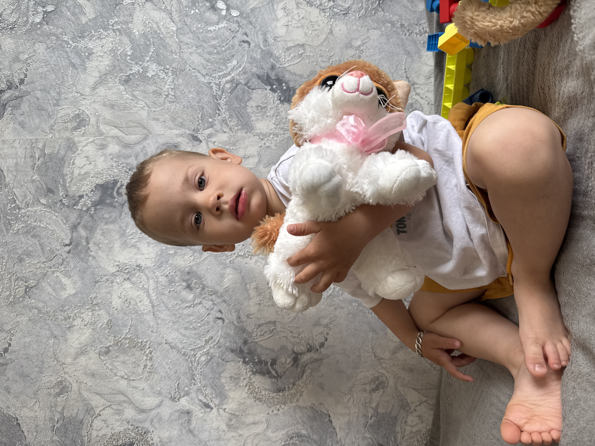 A toddler is lying on a gray textured surface, possibly a bed or couch, in an indoor setting. He is hugging a stuffed animal cat with a pink ribbon. The child is wearing a white t-shirt and yellow shorts, and has a silver bracelet on his left wrist. In the background, there are colorful building blocks and a partially visible stuffed animal bear. The backdrop is a textured gray wall. The location could be in Rusanovo, Russia, or anywhere, since there is no clear indication of location in the image.