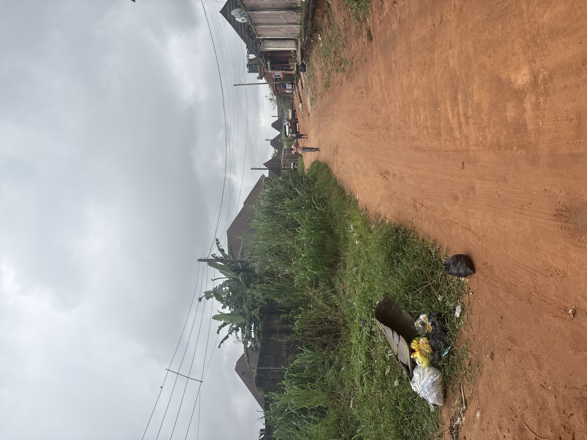 A dirt road in Obosi, Nigeria, is lined with houses and overgrown vegetation. The sky is overcast, with a few scattered clouds. On the left side of the road, a pile of trash, including plastic bags and debris, is accumulated near a patch of tall grass. Further down the road, two women and a child are walking, with a car parked behind them. On the right side of the road, more houses are visible, some with satellite dishes on their roofs. Power lines are strung across the sky, supported by poles.
