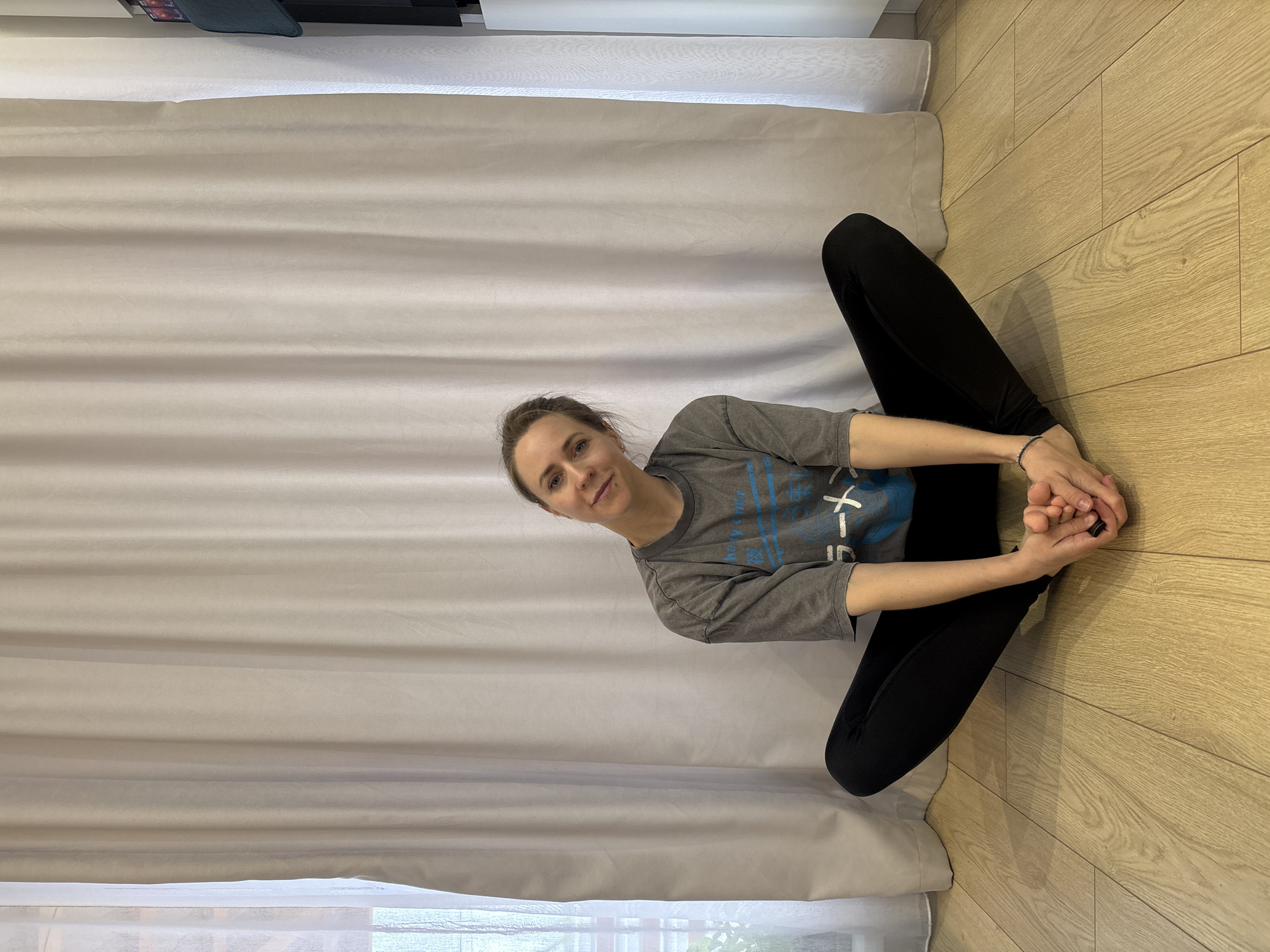 A woman is seated on a wooden floor against a beige curtain, possibly in Chaikovsky, Russia. She is wearing a gray t-shirt with blue lettering and black leggings. Her legs are bent and she is holding her feet. Her face is visible and she is looking towards the camera. A window is visible behind the curtain at the bottom of the image.