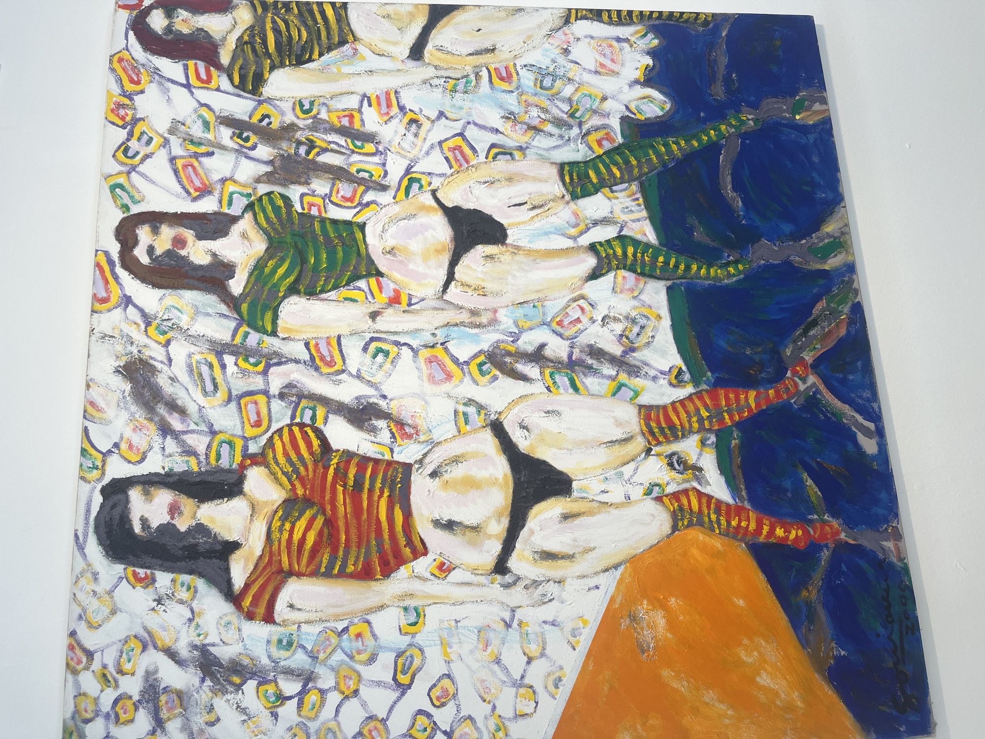The image is a painting. The painting depicts three nude or partially nude figures lying on a surface with a patterned background. The figures are depicted in similar poses, with their backs to the viewer. Each figure has dark hair and is wearing a striped top, though the colors vary. The figures also appear to be wearing bottoms. The surface they are lying on has a white background with multicolored geometric patterns. To the right, a dark blue area appears. An orange triangular form is visible at the bottom. The location is not evident from the image.
