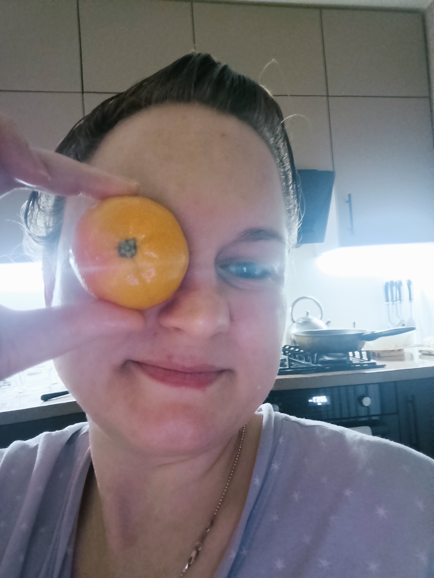 A woman is holding a small, round, orange object, possibly a fruit, in front of her right eye. She is indoors, likely in a kitchen setting, as there are kitchen cabinets, a stove with a pan, and kitchen utensils visible in the background. The woman's expression is neutral, with a slight smile. She is wearing a light-colored, patterned shirt and a thin necklace. The photograph appears to be taken at eye level. The location information Марковский, Russia is provided.