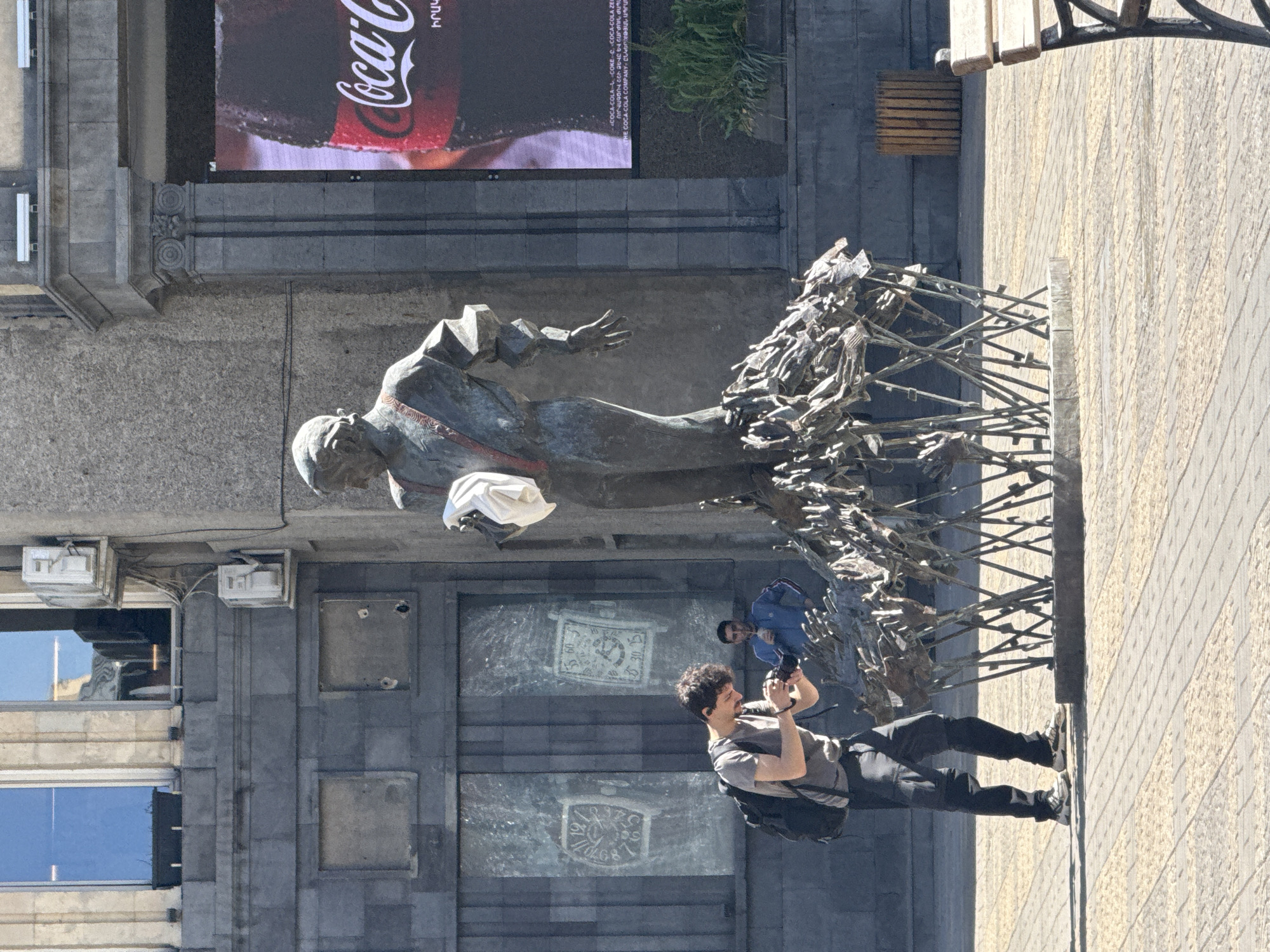 The image depicts a statue of Arno Babajanian in Yerevan, Armenia. A man stands on the sidewalk in front of the statue taking a picture with his phone. A second man is standing close to the statue. Above the statue is an advertisement for Coca-Cola. The buildings in the background are made of gray stone.