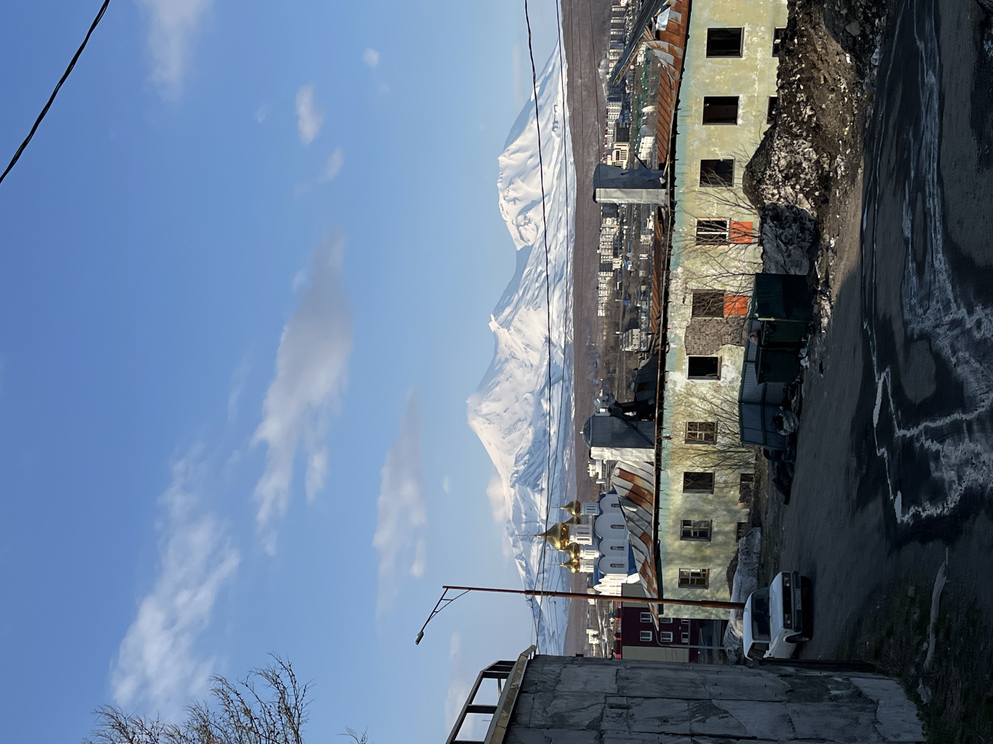 The image presents a view in Petropavlovsk-Kamchatsky, Russia. In the foreground, a dilapidated building under construction stands with exposed steel framework. A rusted corrugated metal roof is visible on a nearby building. A road runs adjacent to a building, with debris and a small white car parked alongside. In the distance, a Russian Orthodox church with golden domes stands out against a backdrop of snow-covered mountains. A clear blue sky is visible above, with scattered clouds. Power lines cut across the upper left of the frame. The overall scene suggests a contrast between urban decay and natural beauty.