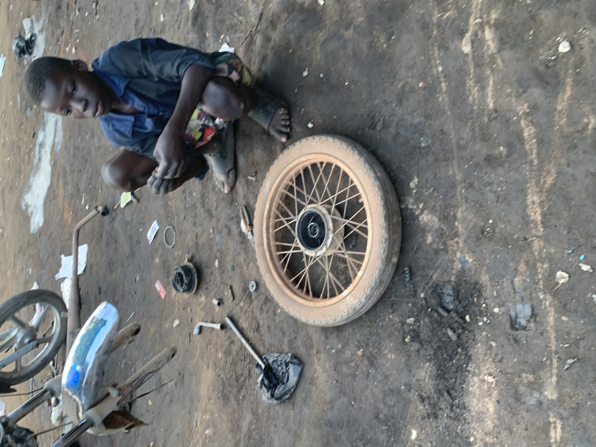 In Shagamu, Nigeria, a young boy is crouched on the dirt ground, focused on disassembling a motorcycle wheel. He wears a dark blue shirt and patterned shorts, with dark sandals on his feet. The wheel, partially disassembled with spokes visible, lies in front of him. Scattered around are tools, including a wrench, and a piece of motorcycle bodywork with blue and white reflective tape, suggesting it might be a fender. The ground is a mix of dirt and debris, with discarded items and a partially visible motorcycle in the background. The scene appears to be an outdoor workshop or repair area, given the presence of motorcycle parts and tools.