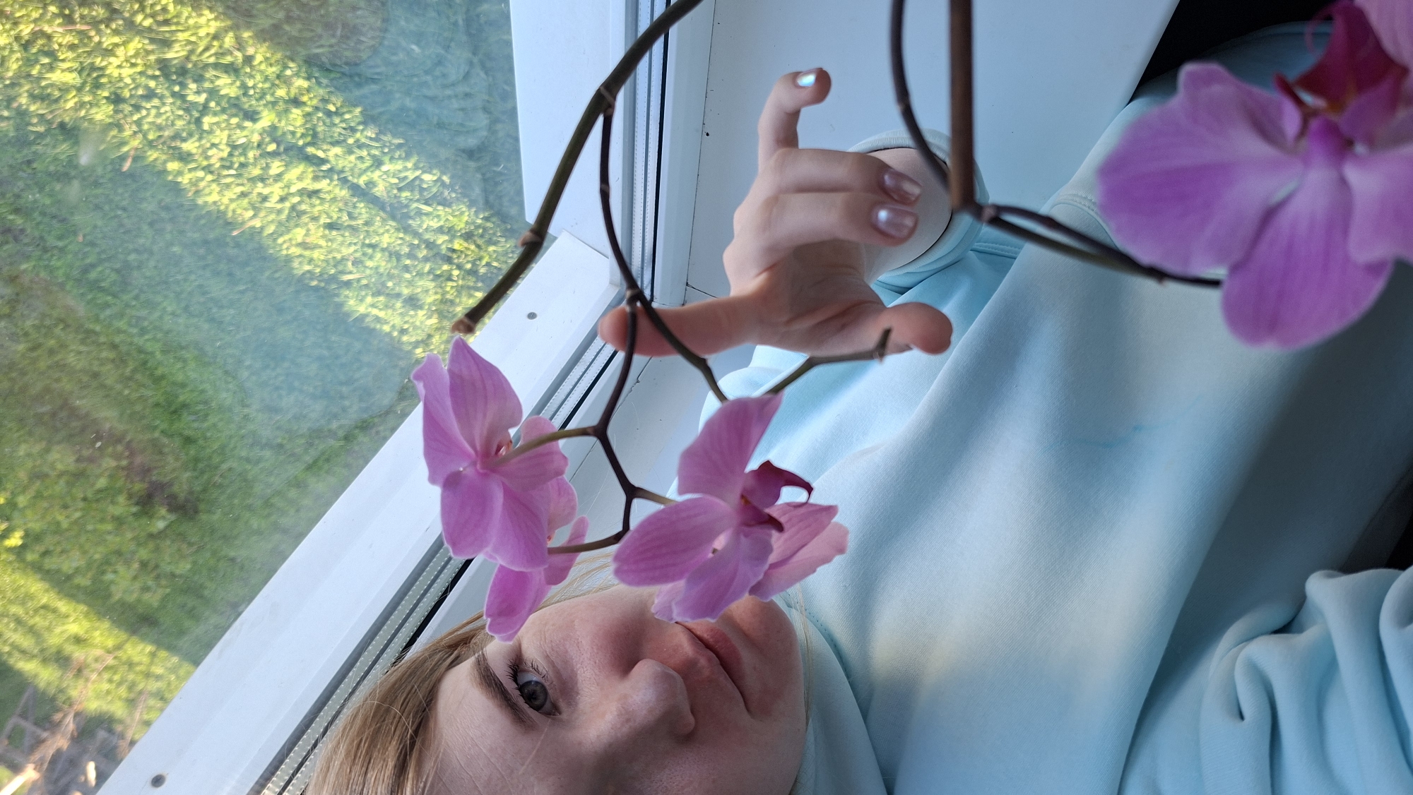 A woman in a light blue hooded sweatshirt is lying down, face near a window. The window overlooks a grassy area, possibly a lawn. In front of the woman's face is a stem of pink orchids. Her hand is raised and positioned near the stem of the orchids. The scene is lit with natural daylight. The location is assumed to be in Большой Букор, Russia.
