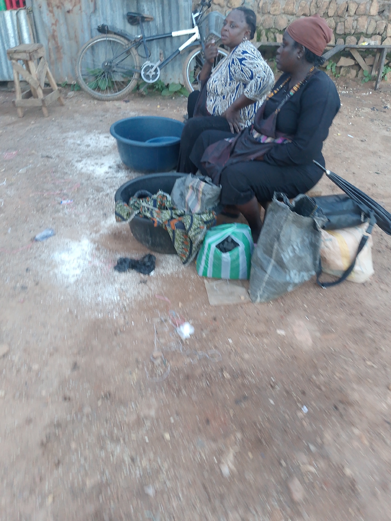 Two women are sitting on the ground in Bukuru, Nigeria, with a bicycle behind them and various objects around them. The woman on the left is wearing a blue and white patterned top and black pants. She has her hand on her lap and is looking to her left. The woman on the right is wearing a brown headwrap, a black top, and black pants. She has a necklace around her neck and is looking to her right.
In front of the women, there are two dark plastic basins. The basin on the left contains what appears to be clothing with a patterned fabric. The basin on the right is empty. To the right of the basins, there are several bags and an umbrella. One bag is green and white striped and appears to contain charcoal. Another bag is dark gray and made of a rough material. There is also a black purse or handbag and an open black umbrella.
The ground is a dirt surface with scattered white powder and debris. Behind the women and basins, a bicycle is leaning against a corrugated metal fence. The bicycle is dark colored with white accents on the handlebars and frame. To the left of the bicycle, a wooden stool with a triangular seat is visible. The background includes a stone wall and some vegetation.