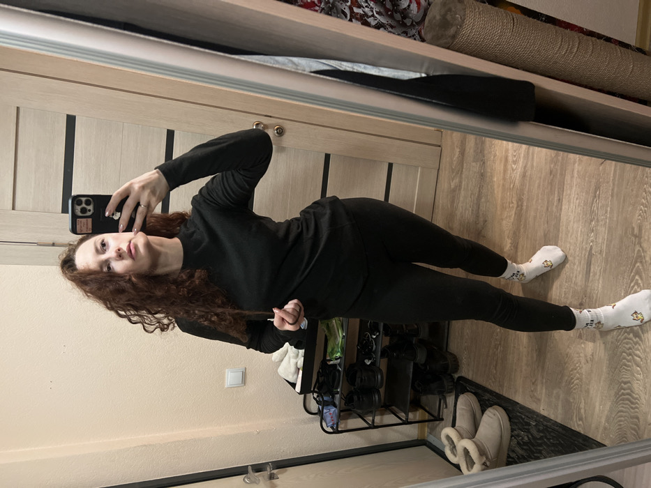 A woman stands in front of a mirror, taking a selfie. She wears a black long-sleeved top and black leggings, along with white socks. Her curly brown hair falls to her shoulders. In the background, there's a door with vertical panels, a shoe rack filled with shoes, and a pair of lined winter boots. Overhead, a storage shelf holds a roll of material and other items. The room has light-colored walls and wooden-look flooring.