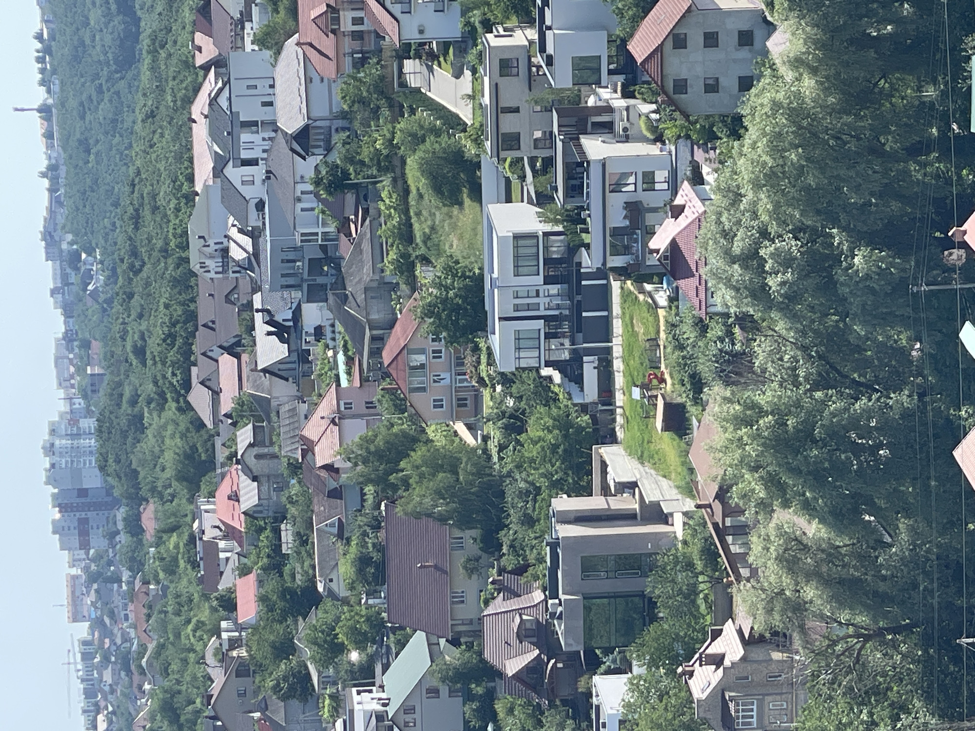 The image is an aerial view of a suburban neighborhood with many houses and trees under a clear sky. The houses are of various sizes and architectural styles, with many featuring red or brown tiled roofs. Some houses are modern, with large windows and flat roofs, while others are more traditional. The neighborhood is densely populated with houses built close to each other. Lush green trees are interspersed throughout the neighborhood, with some areas appearing to be on a slight incline. In the background, taller buildings can be seen, suggesting the presence of a more urban area further away. The overall scene depicts a residential area with a mix of modern and traditional housing surrounded by abundant greenery.