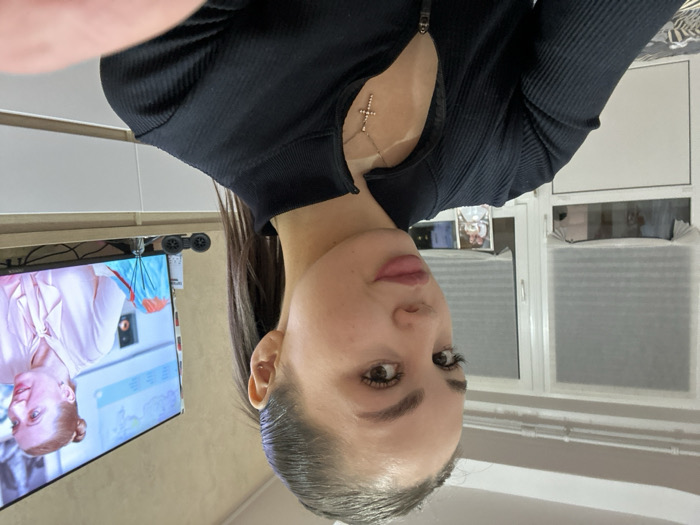 A woman with fair skin and dark hair gazes at the camera. She wears a black, ribbed top that features a plunging neckline adorned with a dainty cross pendant. Behind her, a window covered with blinds can be seen, along with a glimpse of a TV screen displaying a medical drama, reflected in a mirror.