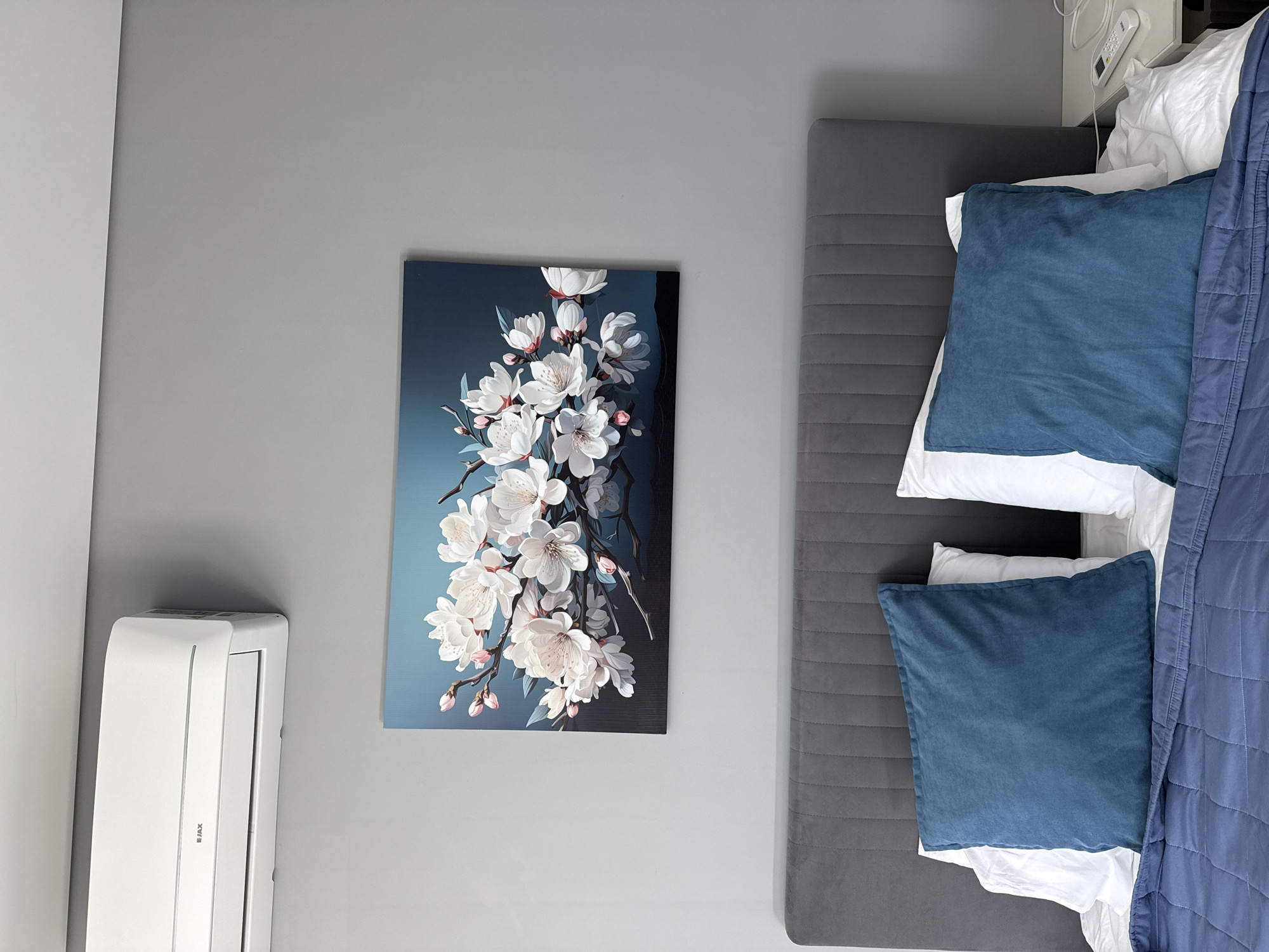 A bedroom interior is depicted in a top-down view. On the right side of the frame, a bed with a grey headboard and base is made up with white sheets, two blue square pillows, and a blue quilted blanket partially covering the bed. To the left of the bed, a grey wall is adorned with a painting of white cherry blossoms against a dark blue background. Further left, a white air conditioning unit is mounted on the wall. The location is Unknown, Unknown. There are no people present.