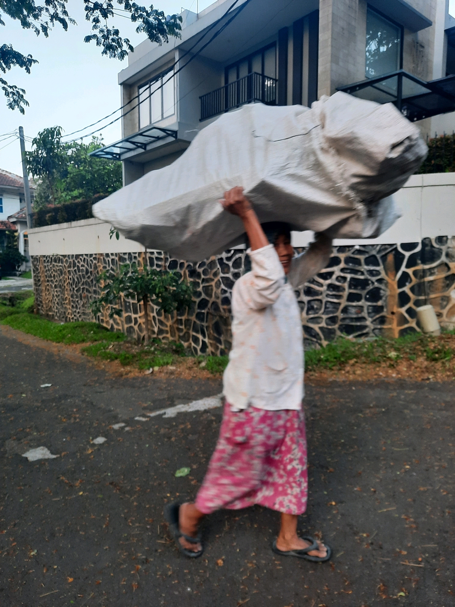 In Babakan Madang, Indonesia, a person walks down a paved road carrying a large, light-colored bundle on their head. The person is wearing a long-sleeved white shirt with a subtle pattern and a vibrant pink floral skirt. They are also wearing dark flip-flops. The bundle appears to be wrapped in a textured material, possibly canvas or plastic sheeting. The background features a modern house with multiple windows and a dark railing on an upper balcony. A textured stone wall, possibly a boundary for the property, runs alongside the road, interspersed with patches of green grass and a small tree. The sky is overcast, suggesting it might be early morning or late afternoon. The road surface is asphalt, with some scattered debris, including leaves and small stones.