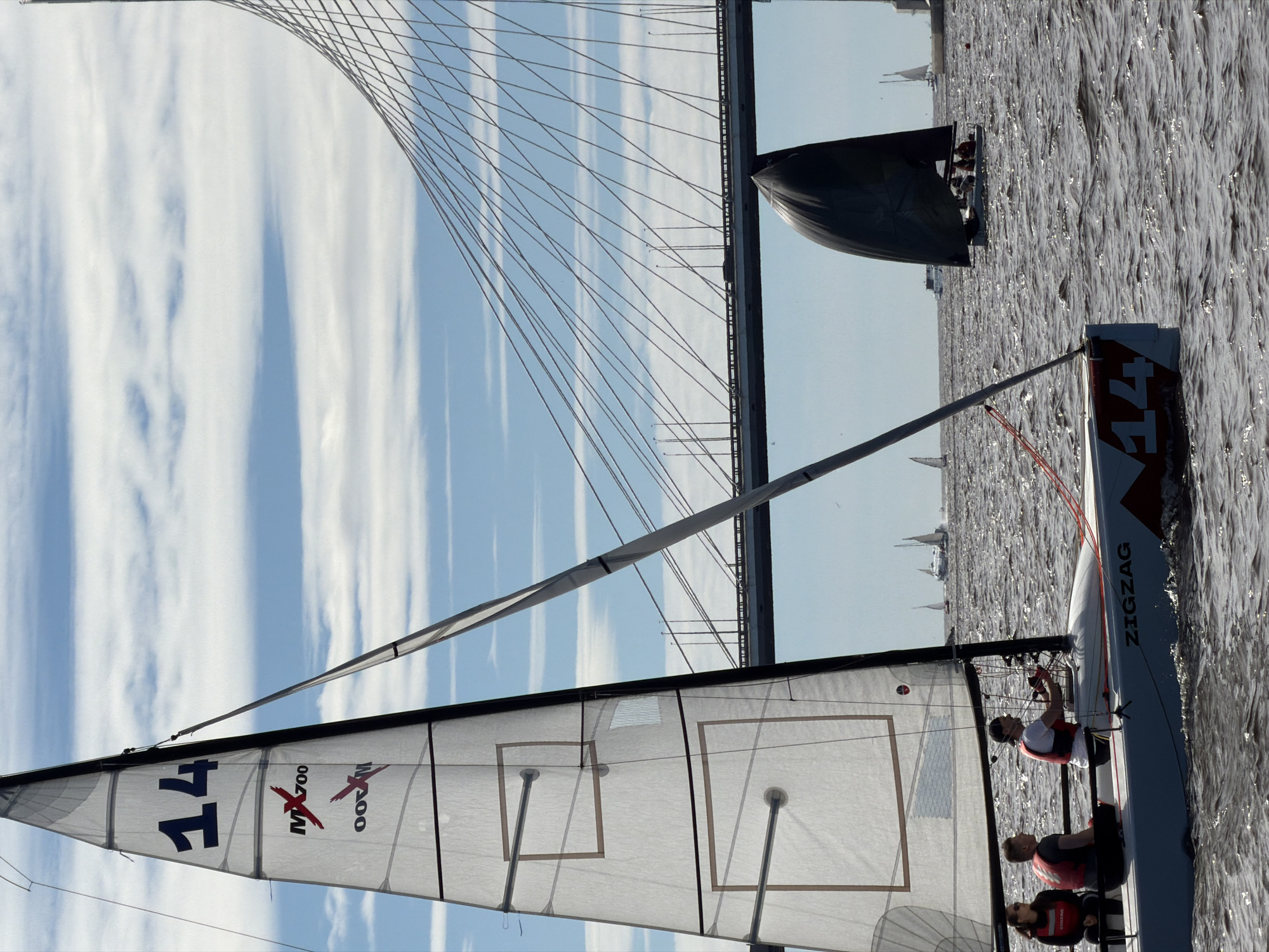 The image depicts a sailing race on the Neva River in Saint Petersburg, Russia, with the cable-stayed bridge, likely the Western High-Speed Diameter bridge, serving as a prominent backdrop. In the foreground, a white sailboat, identified by the number "14" on its sail and the name "ZIGZAG" on its hull, carries three individuals. Two individuals are seated on the port side, one wearing a red life vest, and the other is steering the boat from the starboard side, also in a red life vest and a cap. The sail is translucent, revealing a darker logo and the number "14" in dark blue. The water is choppy and reflects the sunlight. In the middle ground, several other sailboats are visible, one with a dark sail. The bridge spans the river, with traffic visible on its roadway and lamps lining its structure. The sky is blue with scattered white clouds.